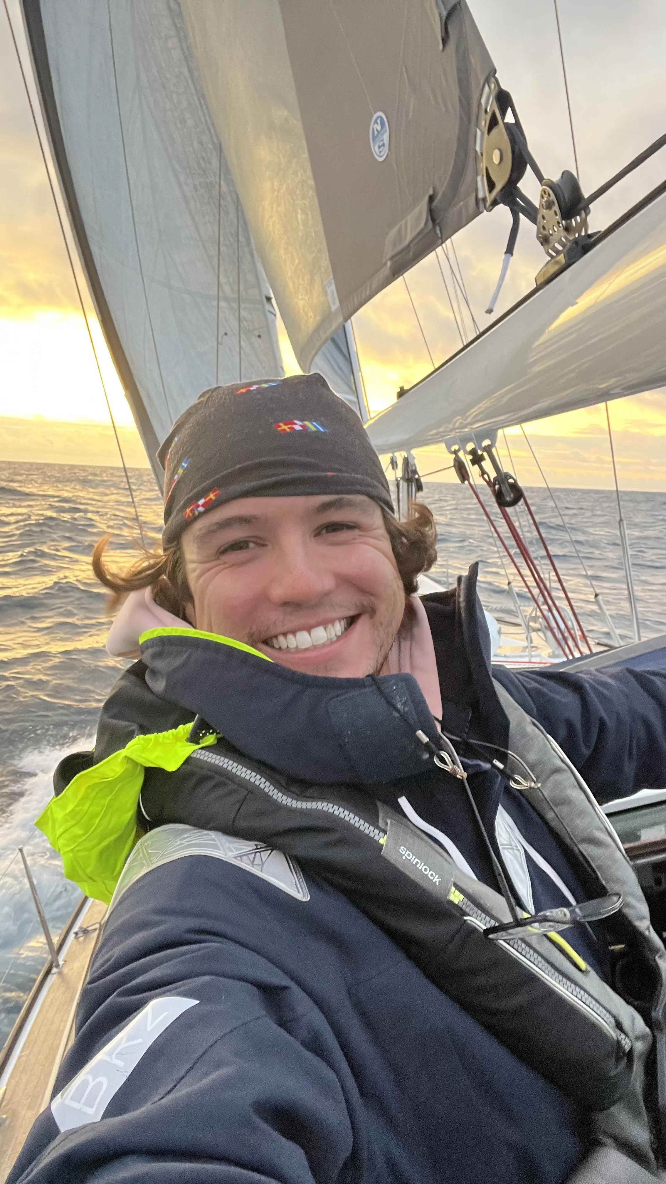 A smiling man taking a selfie on a sailboat during sunset, with sails and ocean waves in the background.