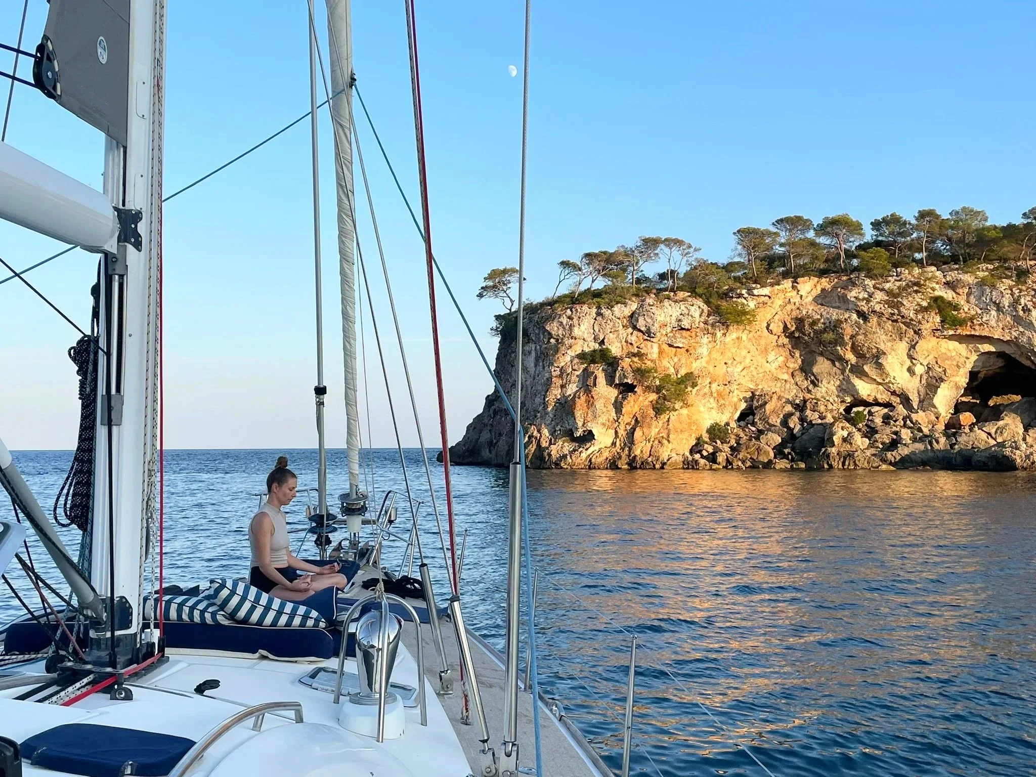 A woman on a sailboat working on a laptop, with a rocky cliff and trees in the background, under a clear blue sky.