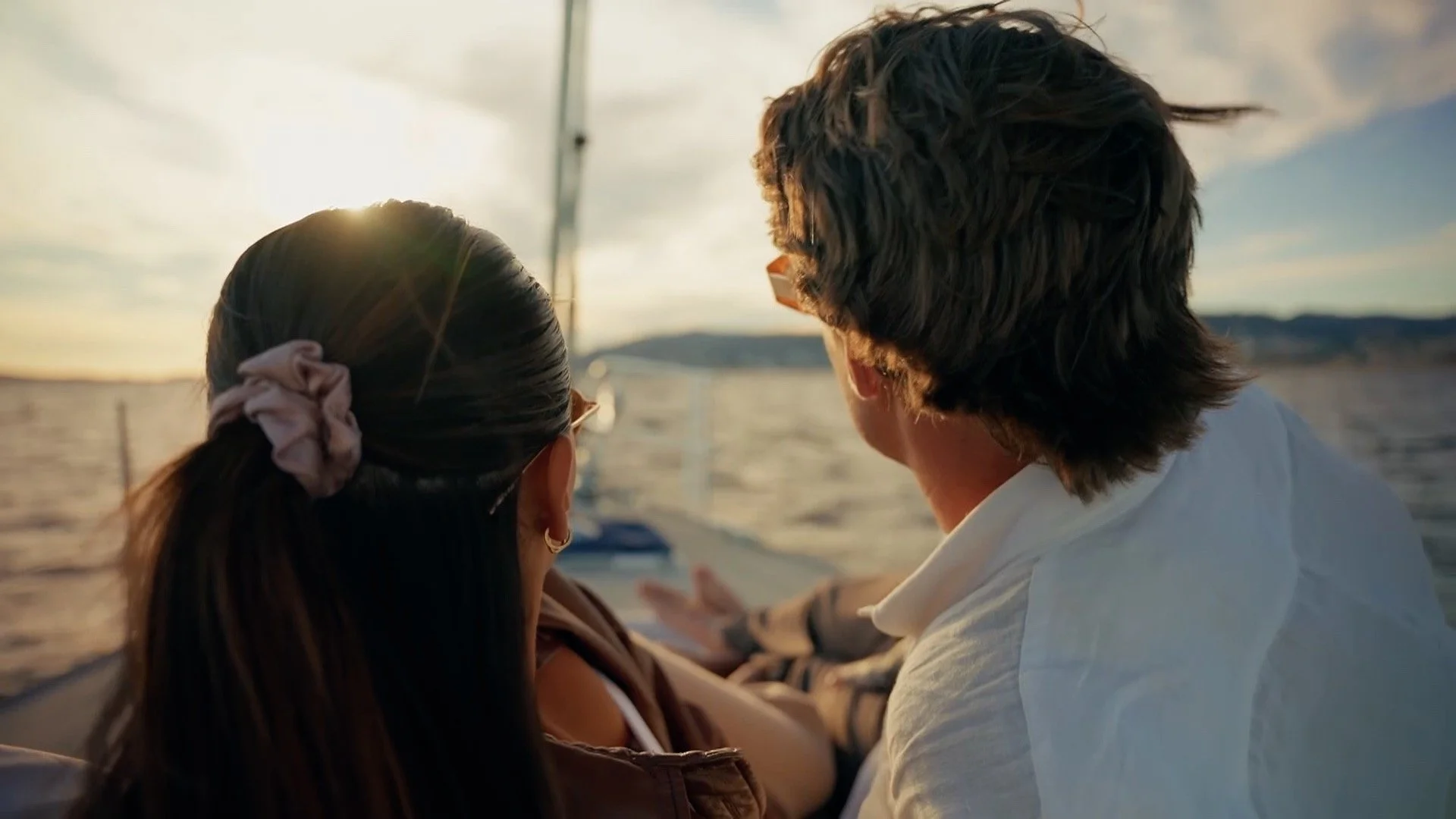 A couple sitting on a boat at sunset, facing a lake with distant hills, enjoying the view.
