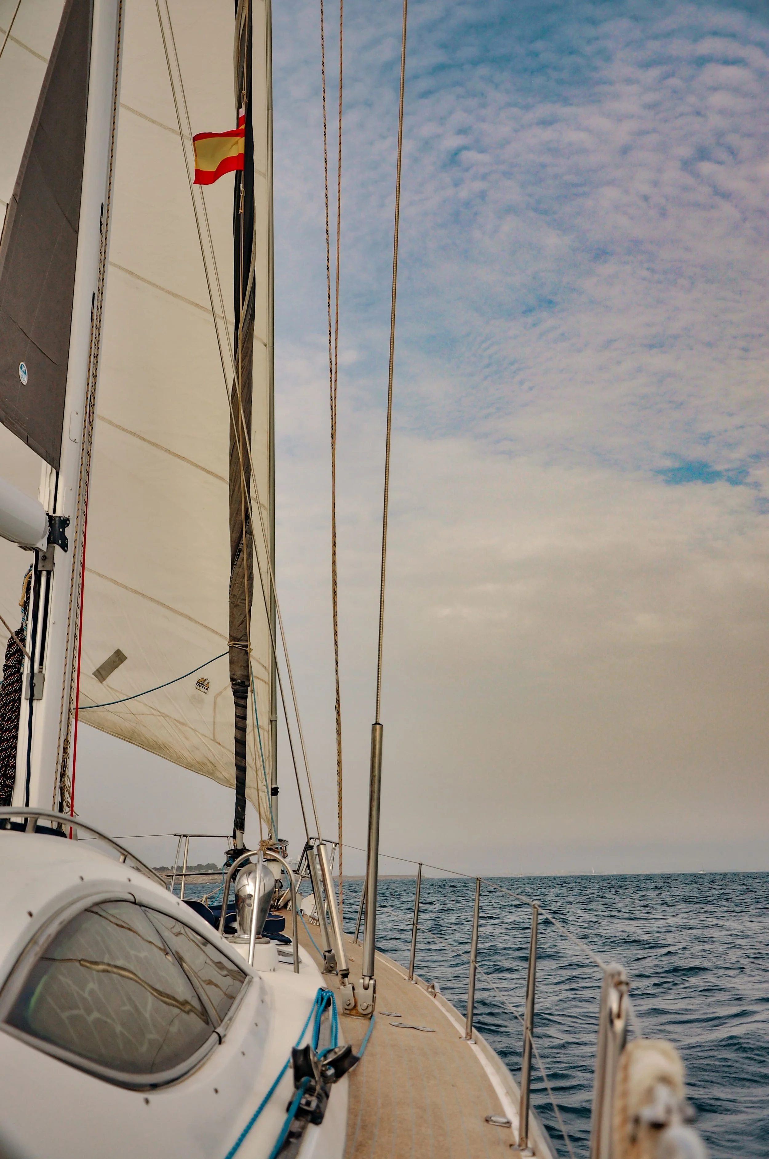 View from a sailboat showing the side deck, part of the sail and rigging, with the open sea and a partly cloudy sky in the background.