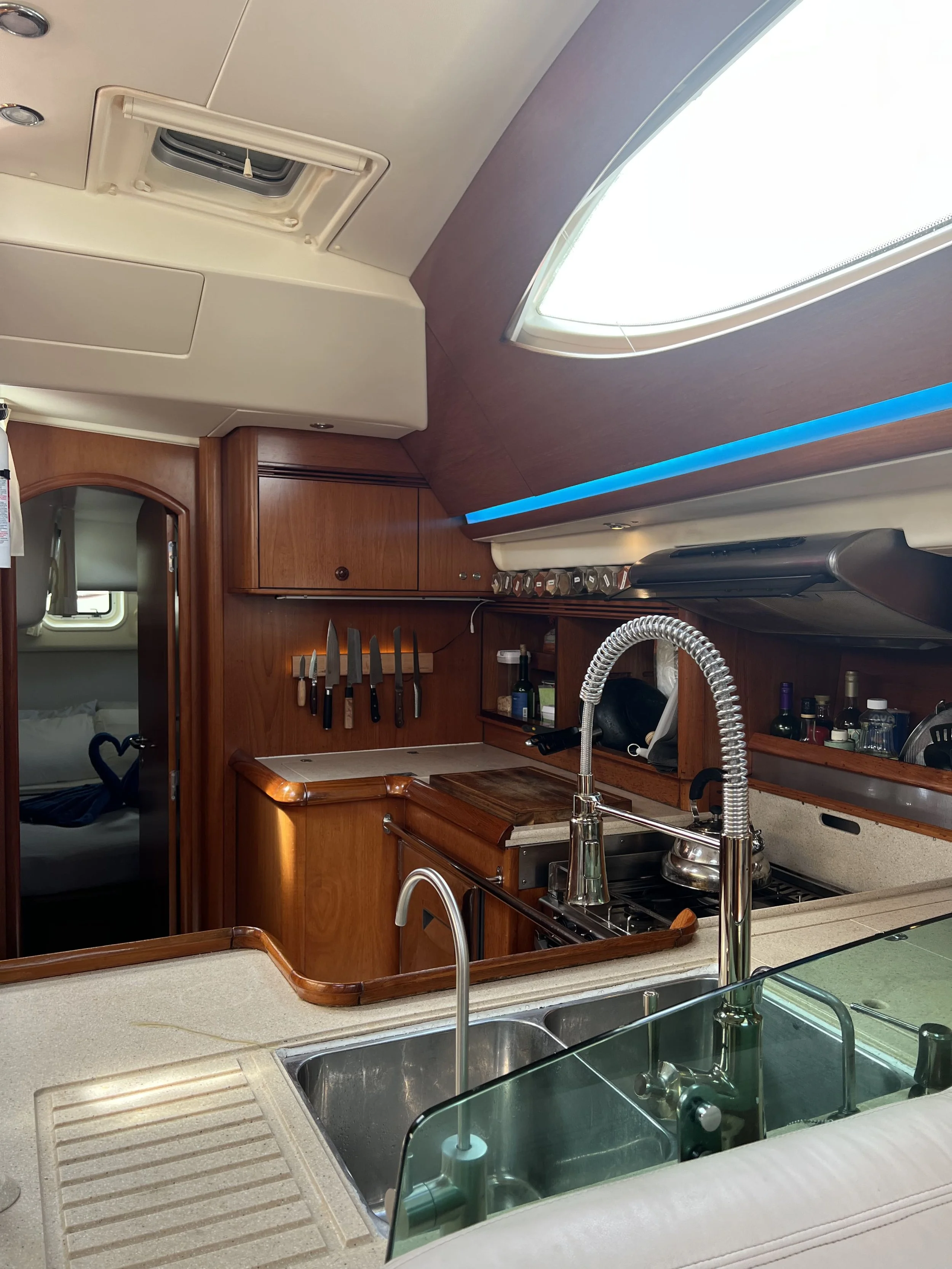 Interior of a boat kitchen with wooden cabinets, a sink, a stovetop with a kettle, and a view of a bedroom through an open door, in a well-lit space with a skylight.