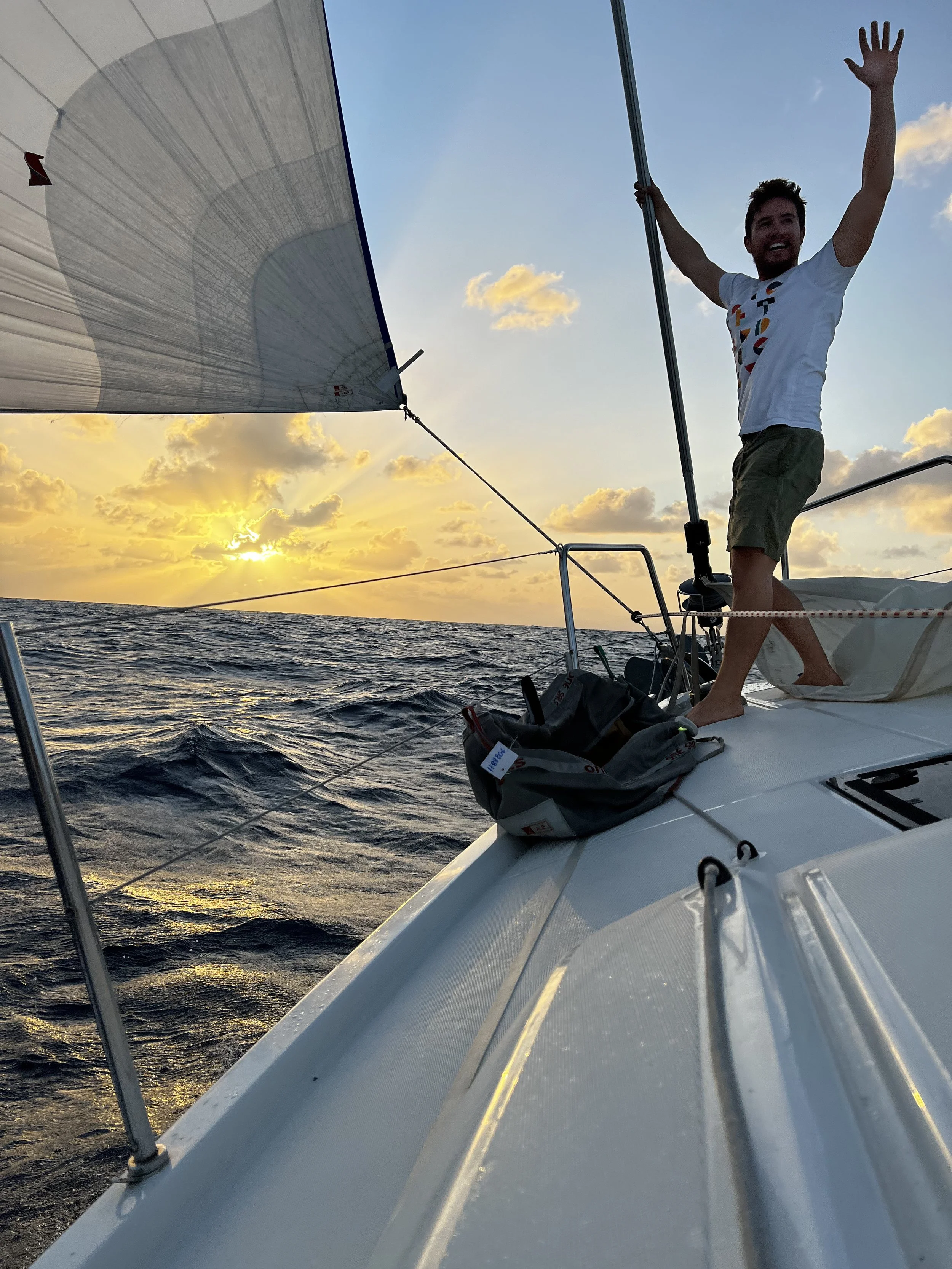A man standing on the deck of a sailboat during sunset, smiling and waving, with the ocean and sky with clouds in the background.