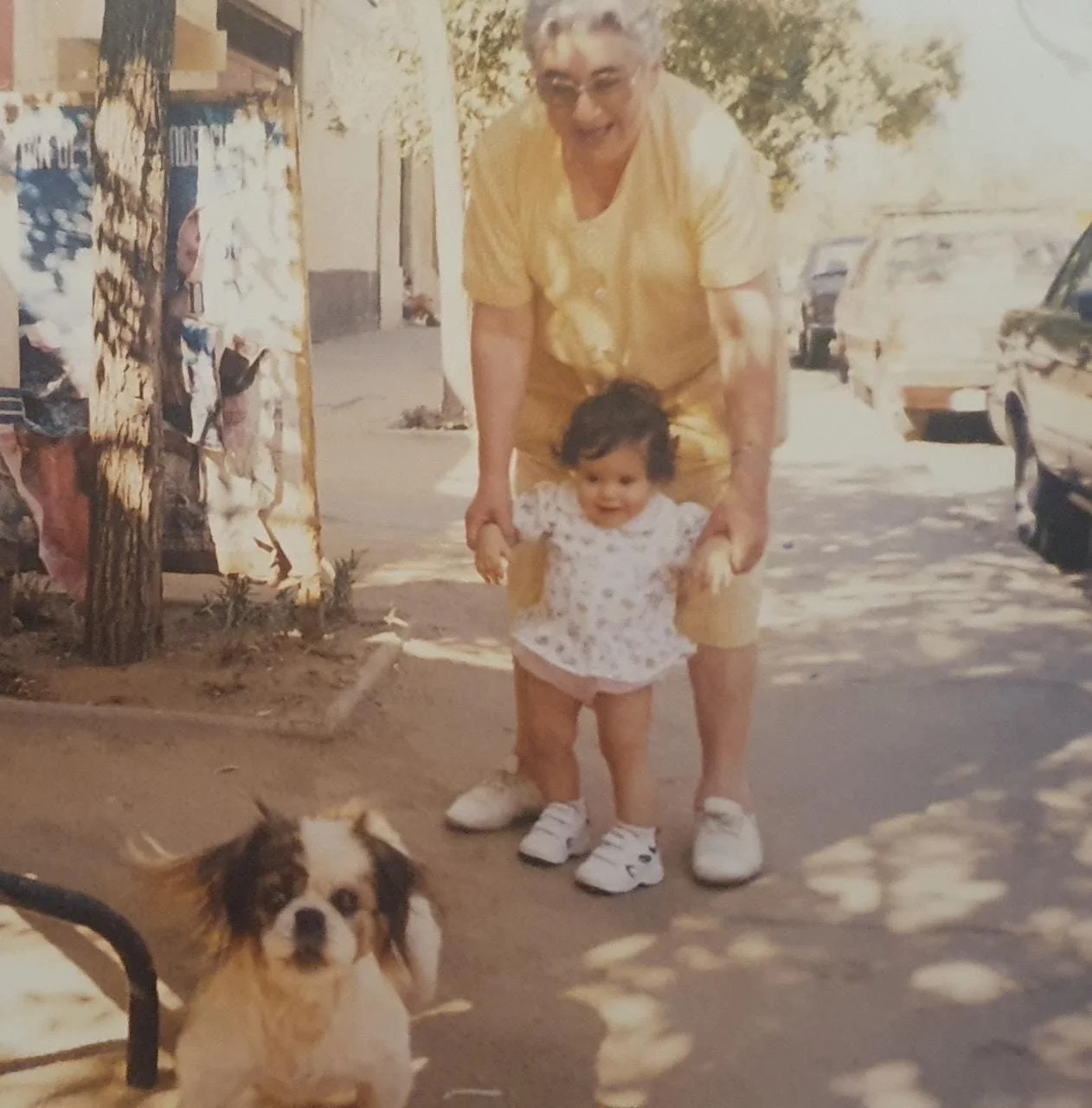 A woman and a young girl holding hands outside on a sunny day, with a dog sitting in front of them, and parked cars in the background.