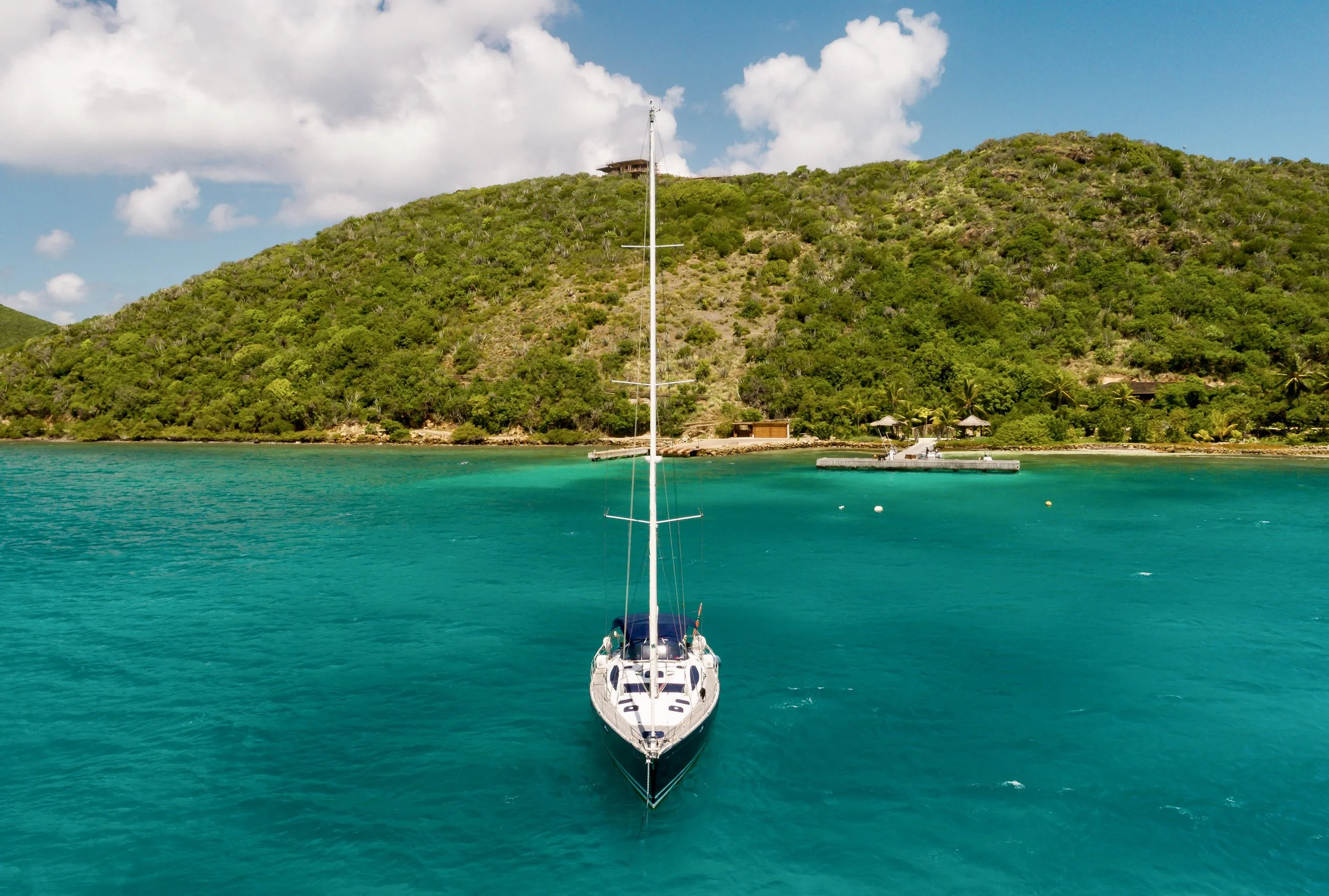 A sailboat floating in turquoise water near a lush green island with hills, buildings, and a small dock.