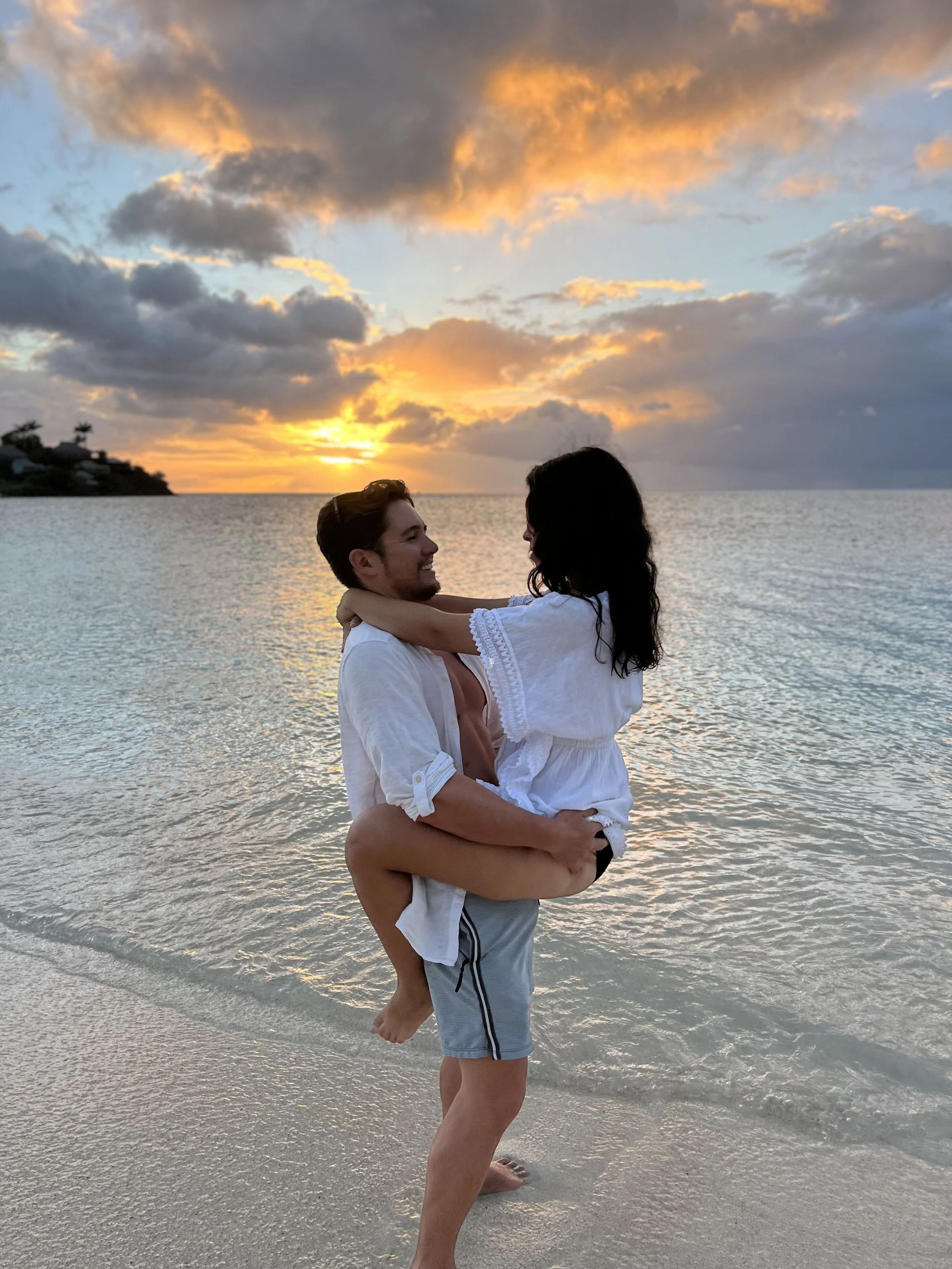 A man lifting a woman on the beach during sunset, with colorful clouds and the ocean in the background.