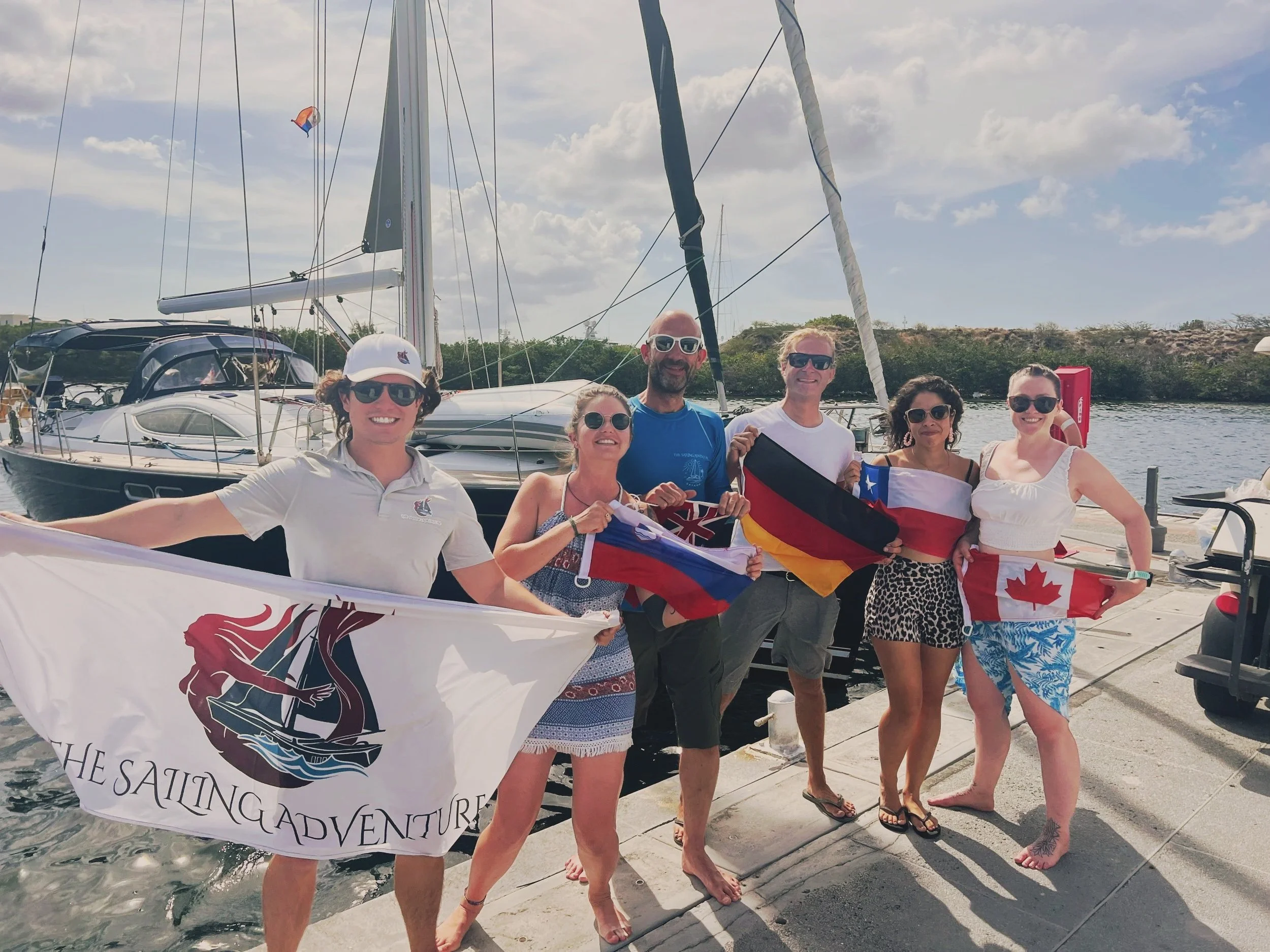 Group of six smiling people holding flags, standing on a dock with boats in the background, under a partly cloudy sky.