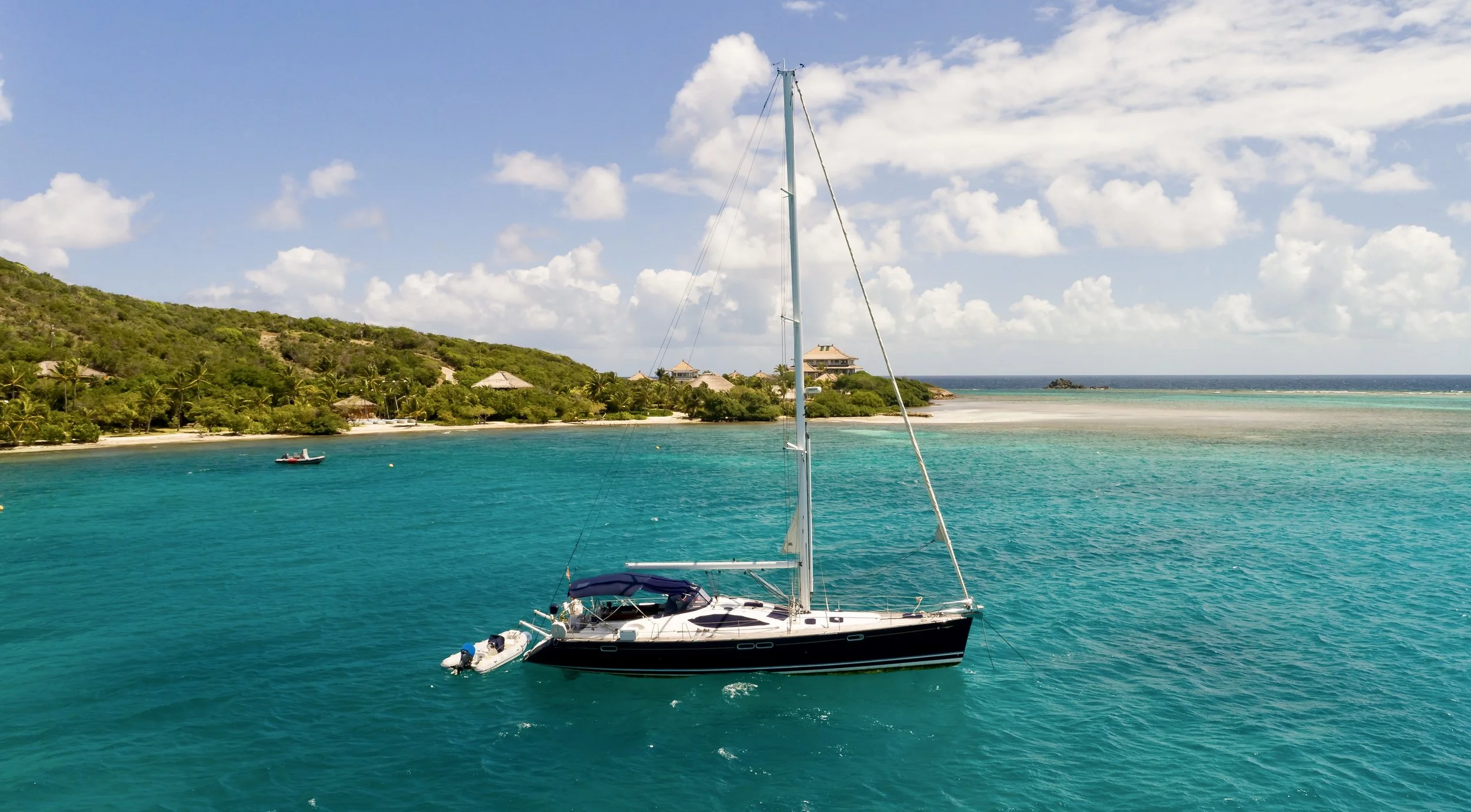 A sailboat anchored near a lush green coastline with trees and houses, under a partly cloudy sky. The turquoise water surrounds the boat, with a small motorboat nearby.