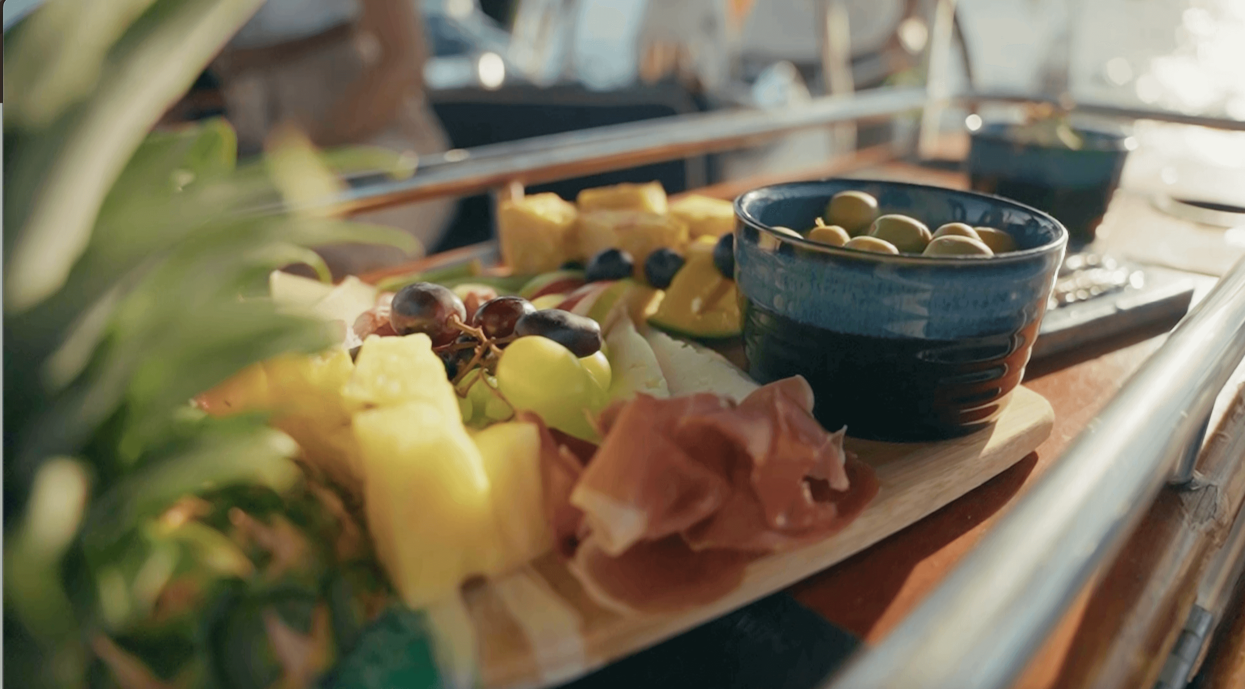 A variety of fresh fruits, green olives in a black bowl, and sliced prosciutto on a wooden serving board, set on a table with a rustic background.