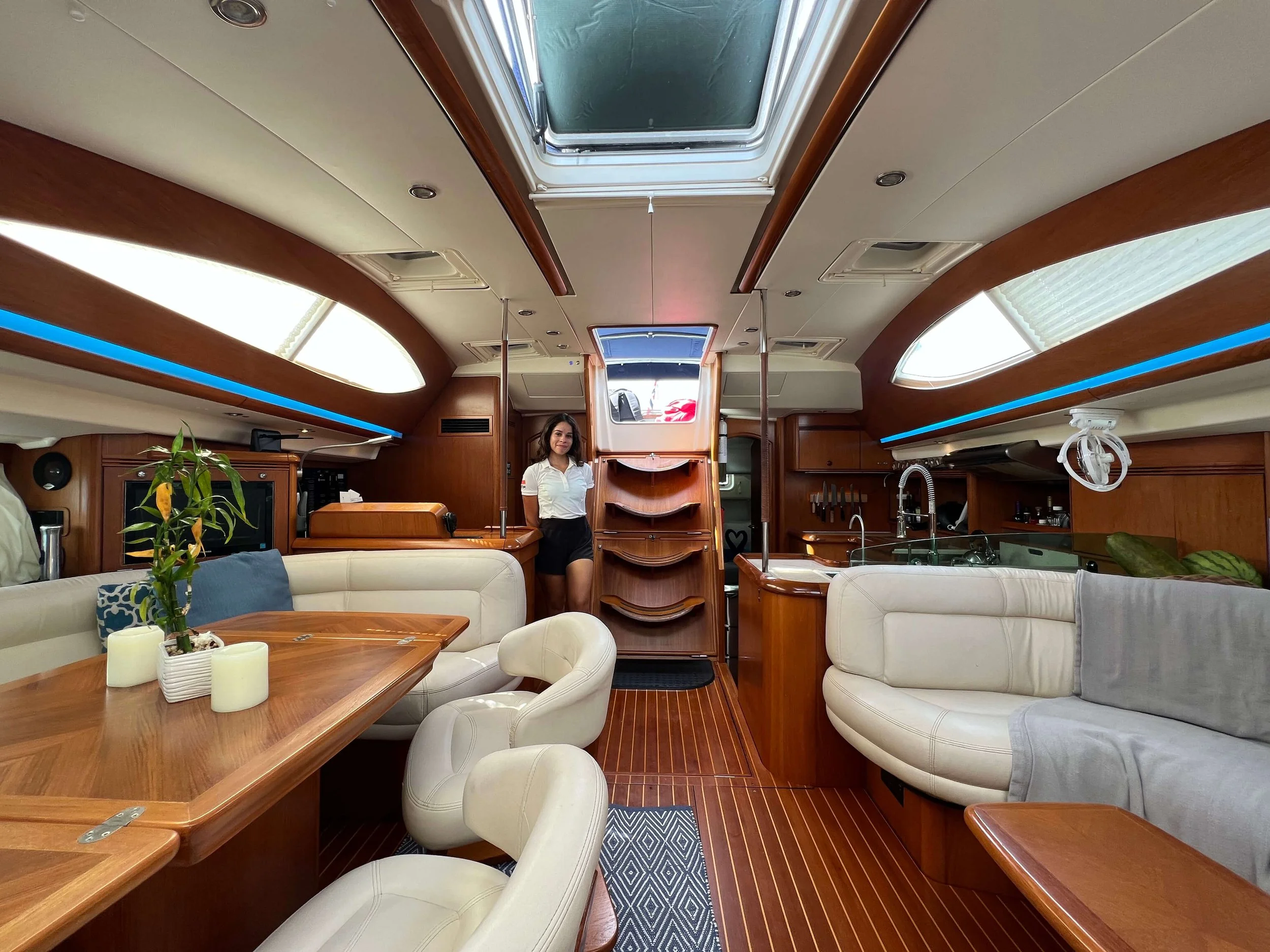 Interior of a yacht with wooden paneling, cream-colored couches, a dining table with candles and a plant, and large windows and skylights letting in natural light. A woman in a white shirt and black shorts stands near a staircase.