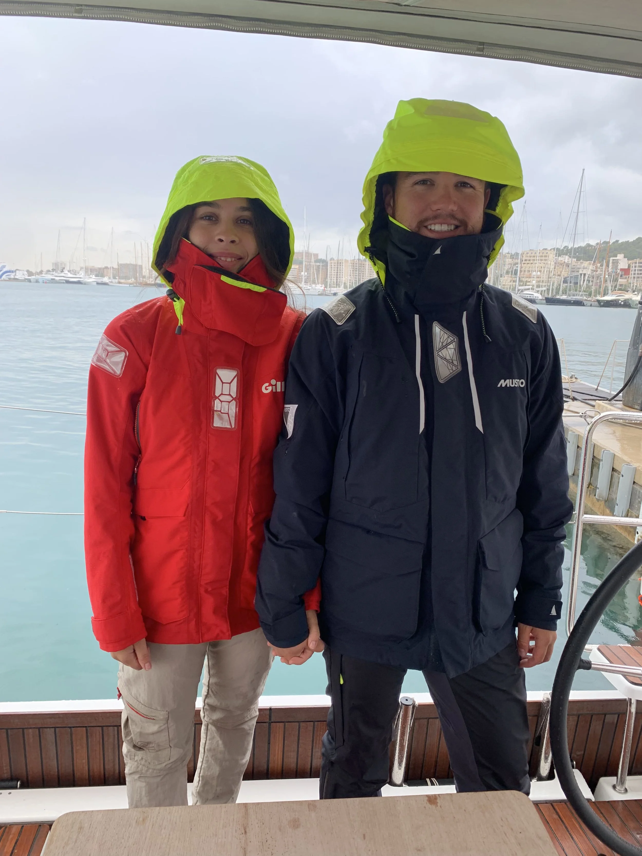 Two people dressed in yellow rain hoods and waterproof jackets, standing on a boat dock, holding hands, with a marina and cloudy sky in the background.