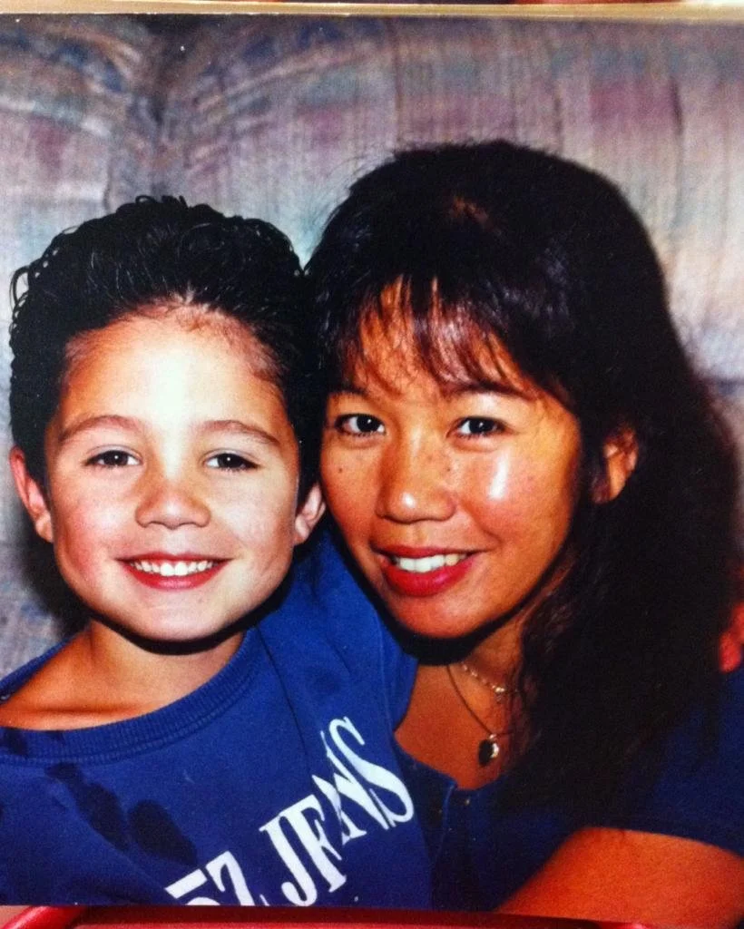 A young boy and woman smiling and posing together indoors, close together with their heads touching. The boy has short, dark, curly hair and is wearing a blue T-shirt. The woman has long, dark hair with bangs, and is wearing a dark shirt with a necklace. They appear happy and affectionate.