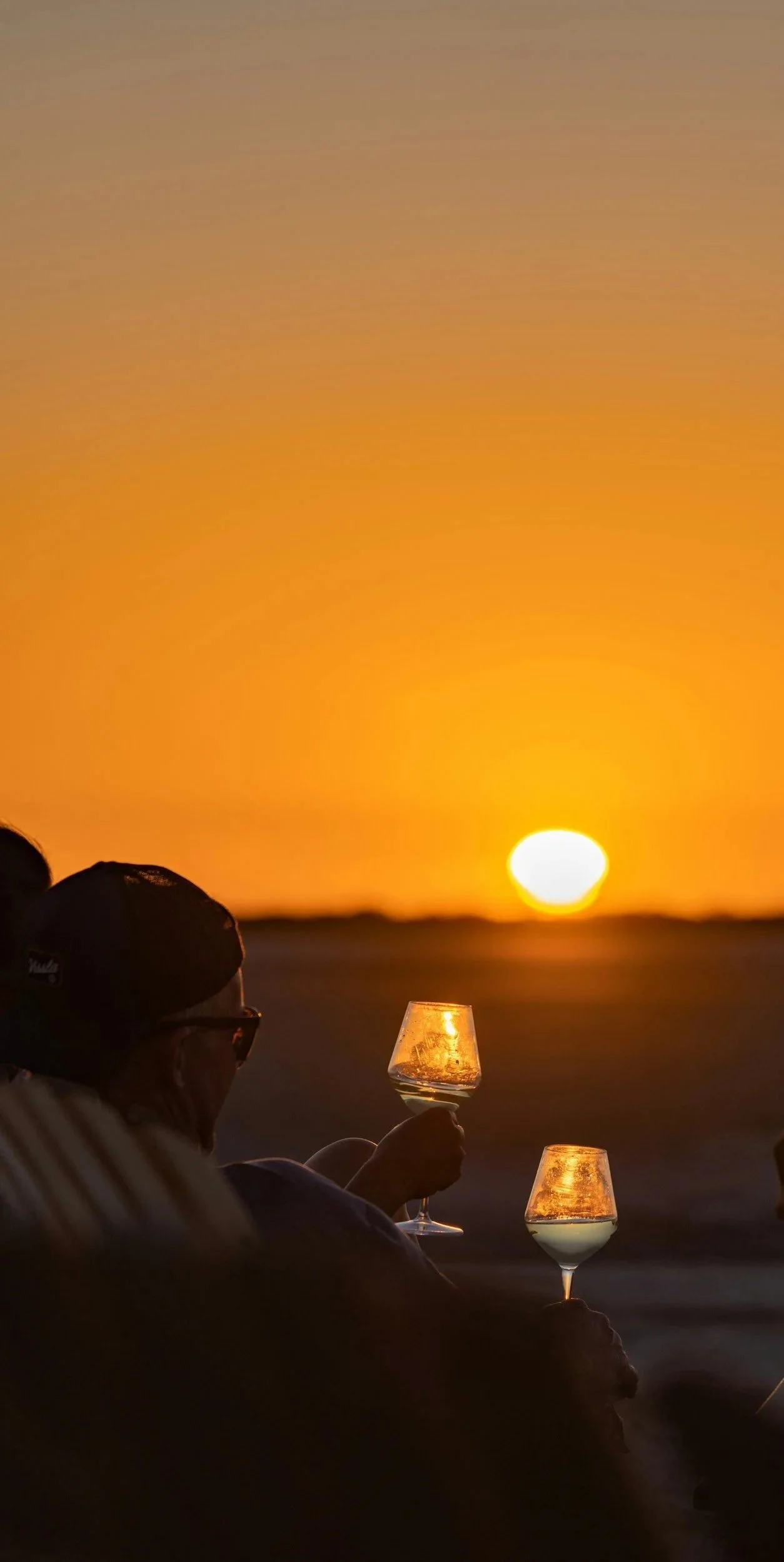 People sitting at the beach during sunset, holding wine glasses.