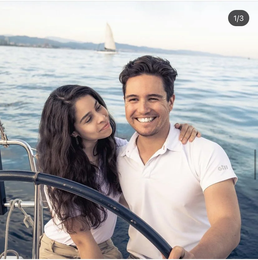 A happy couple smiling on a boat, with a body of water and a sailboat in the background.