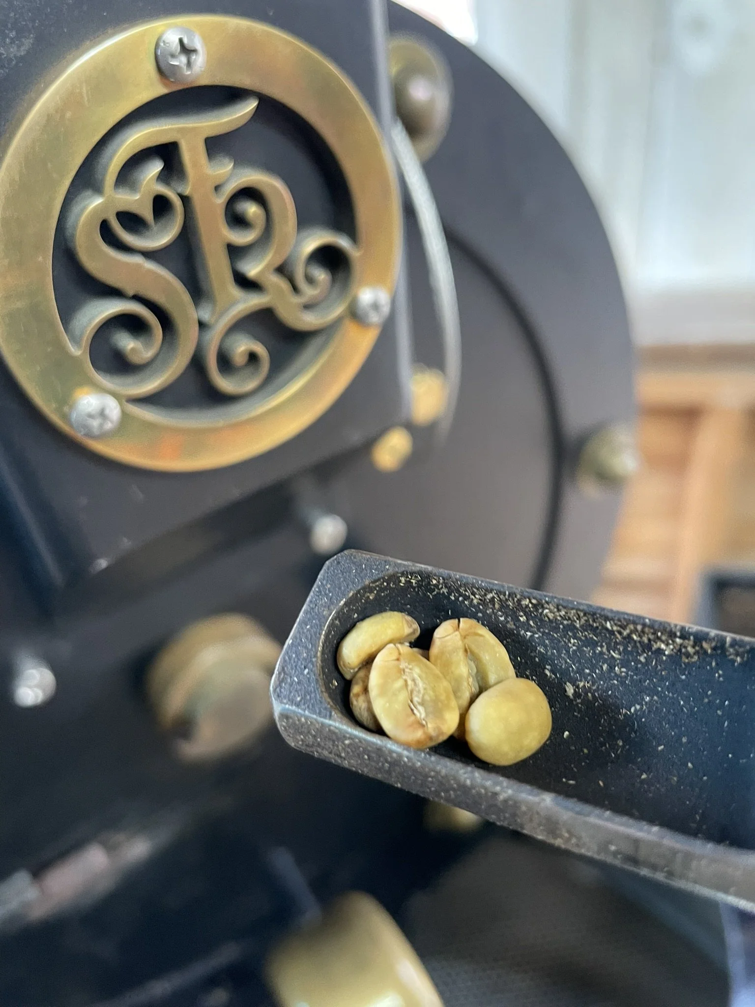 Close-up of coffee beans in a black scoop in front of a vintage coffee grinder with ornate gold and silver details.
