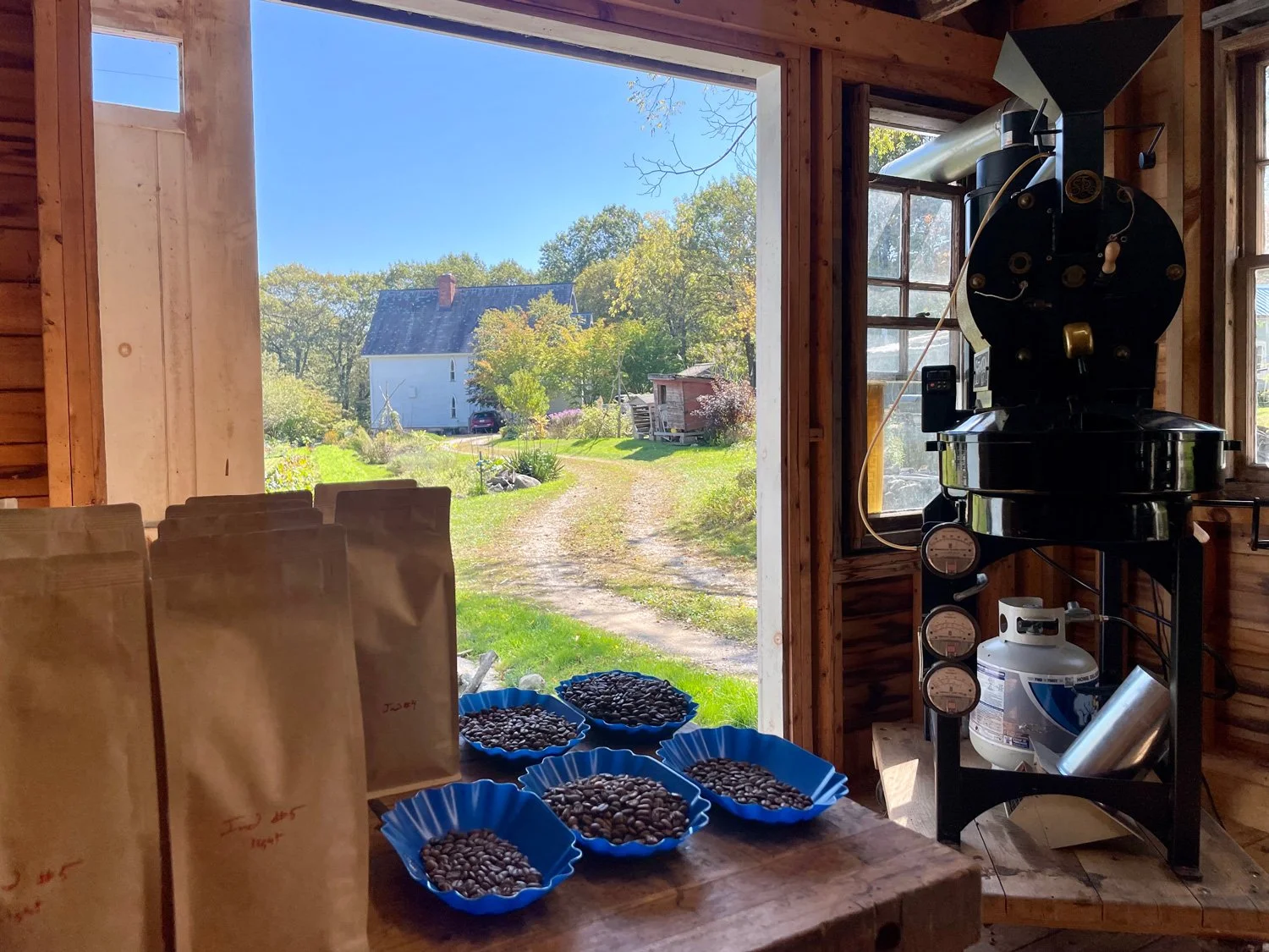 Inside a rustic wooden building, a coffee roaster machine is visible near a window. Plates filled with roasted coffee beans are on a wooden table, with paper bags labeled for coffee storage, overlooking a farm scene with a white house surrounded by trees.