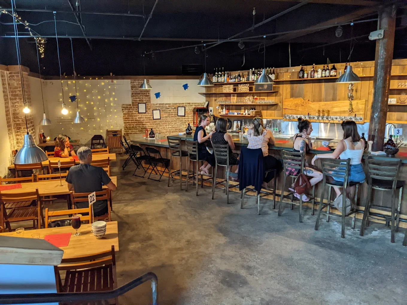 Interior of a cozy bar or restaurant with dim lighting, wooden decor, and a brick wall. Several women are seated at the bar, while a man sits at a table. Hanging pendant lights illuminate the space, decorated with some string lights and wall art.