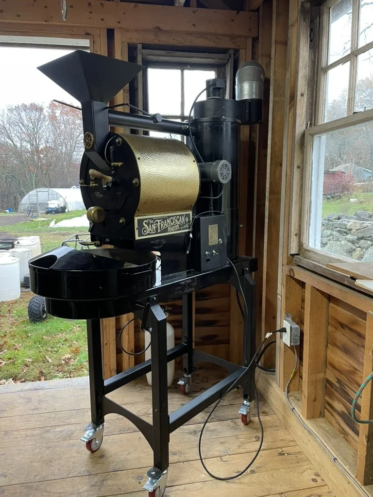 A vintage San Francisco coffee roaster machine inside a wooden room near a window, with outdoor trees and a garden visible outside.