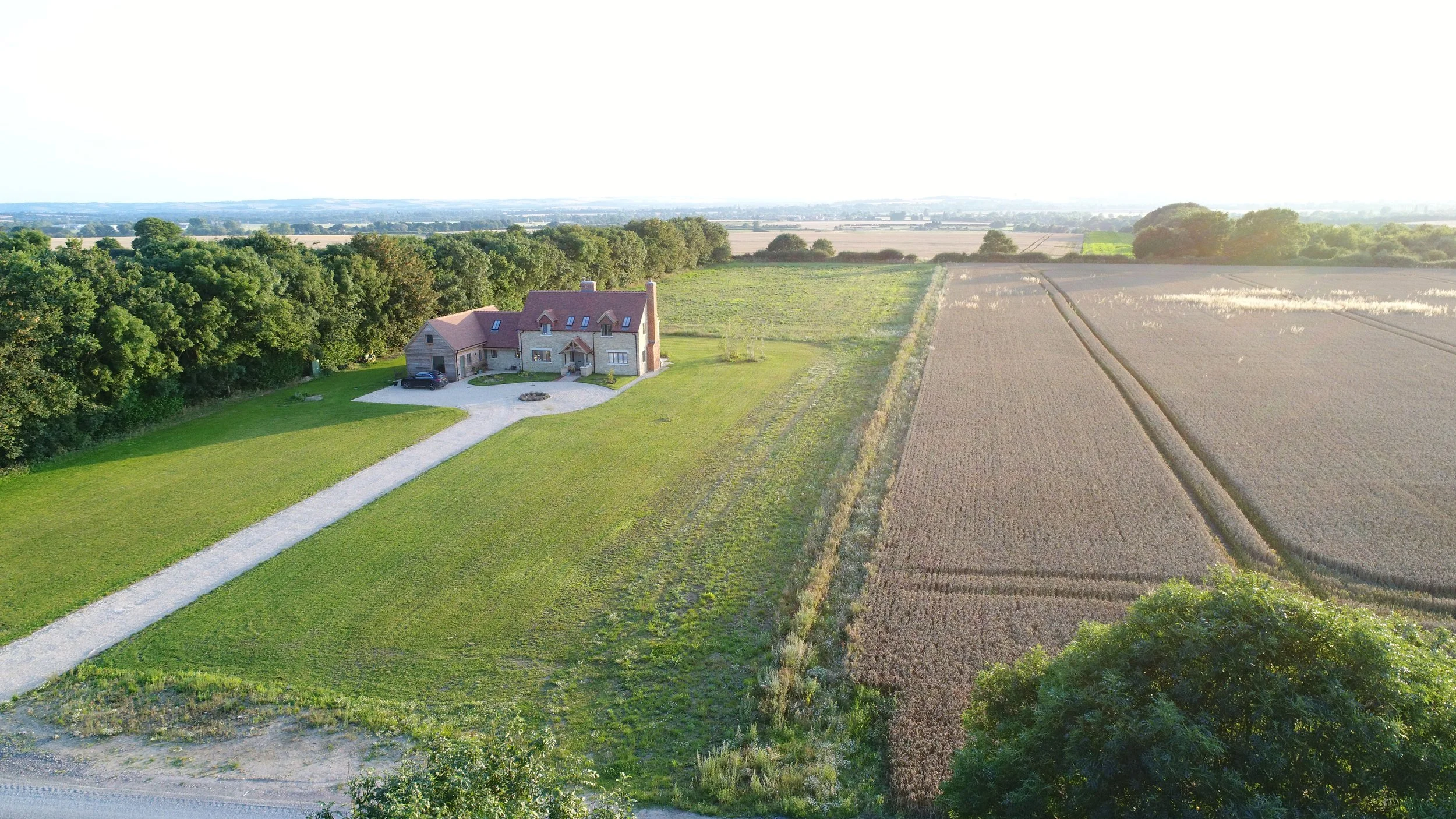 Aerial view of a large two-story house with a driveway, surrounded by green lawn and trees, next to farmland with fields and crop lines, under a bright sky.