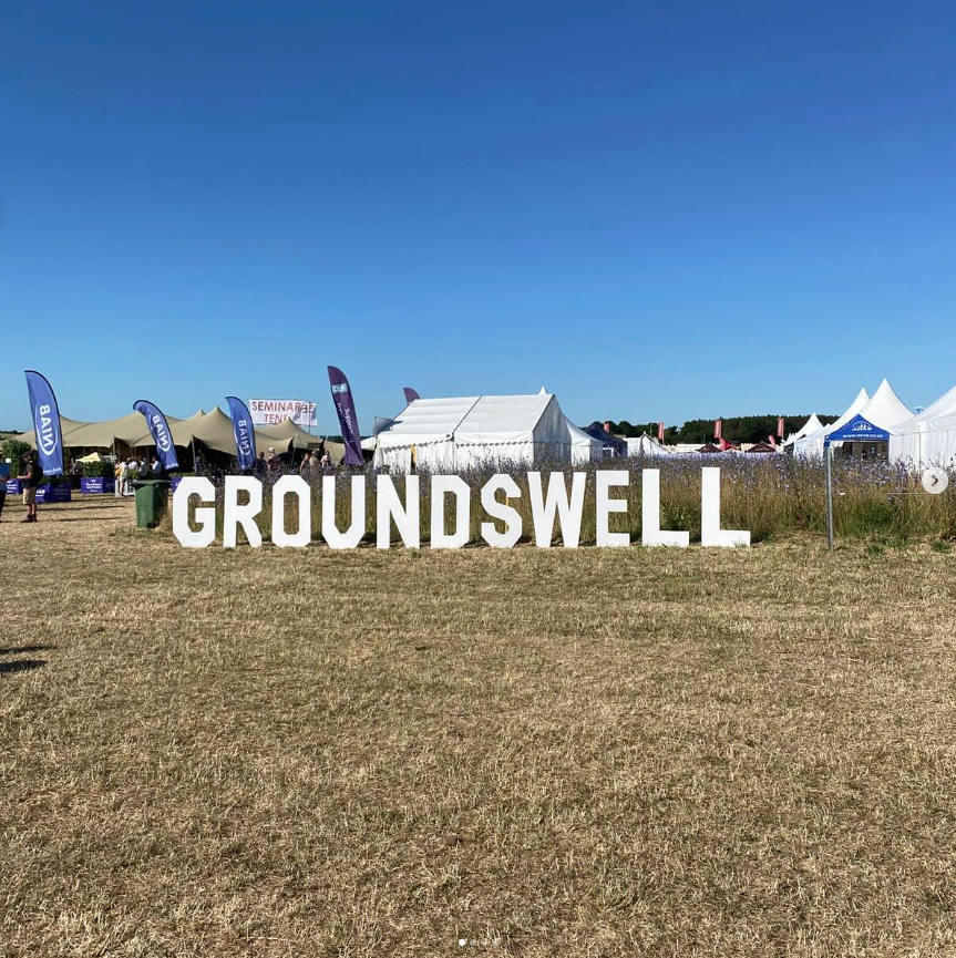 Outdoor festival scene with large white 'GROUNDSWELL' sign, tents, flags, and people on a grassy field under a clear blue sky.