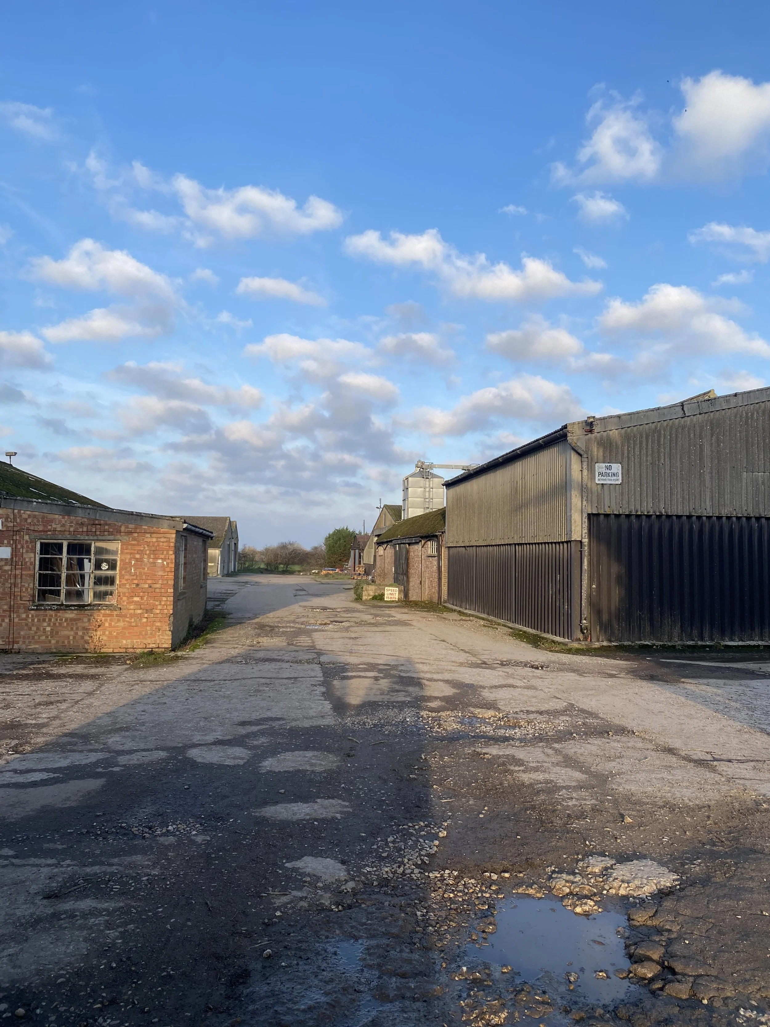 A rural or industrial gravel road with patches of asphalt, puddles, and old buildings on either side, under a partly cloudy blue sky.