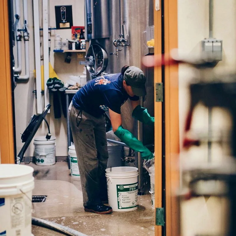A person working in a brewery or industrial setting, wearing green rubber gloves and a baseball cap, handling a bucket.