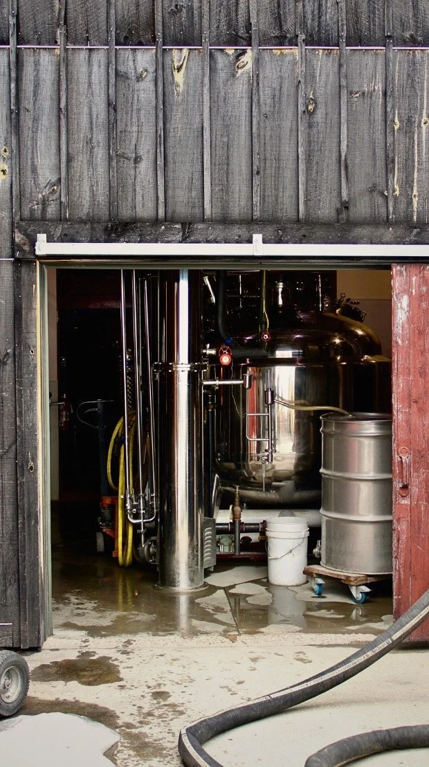 Industrial brewing equipment inside a small brewery or brewery shed, including large stainless steel tanks and pipes.