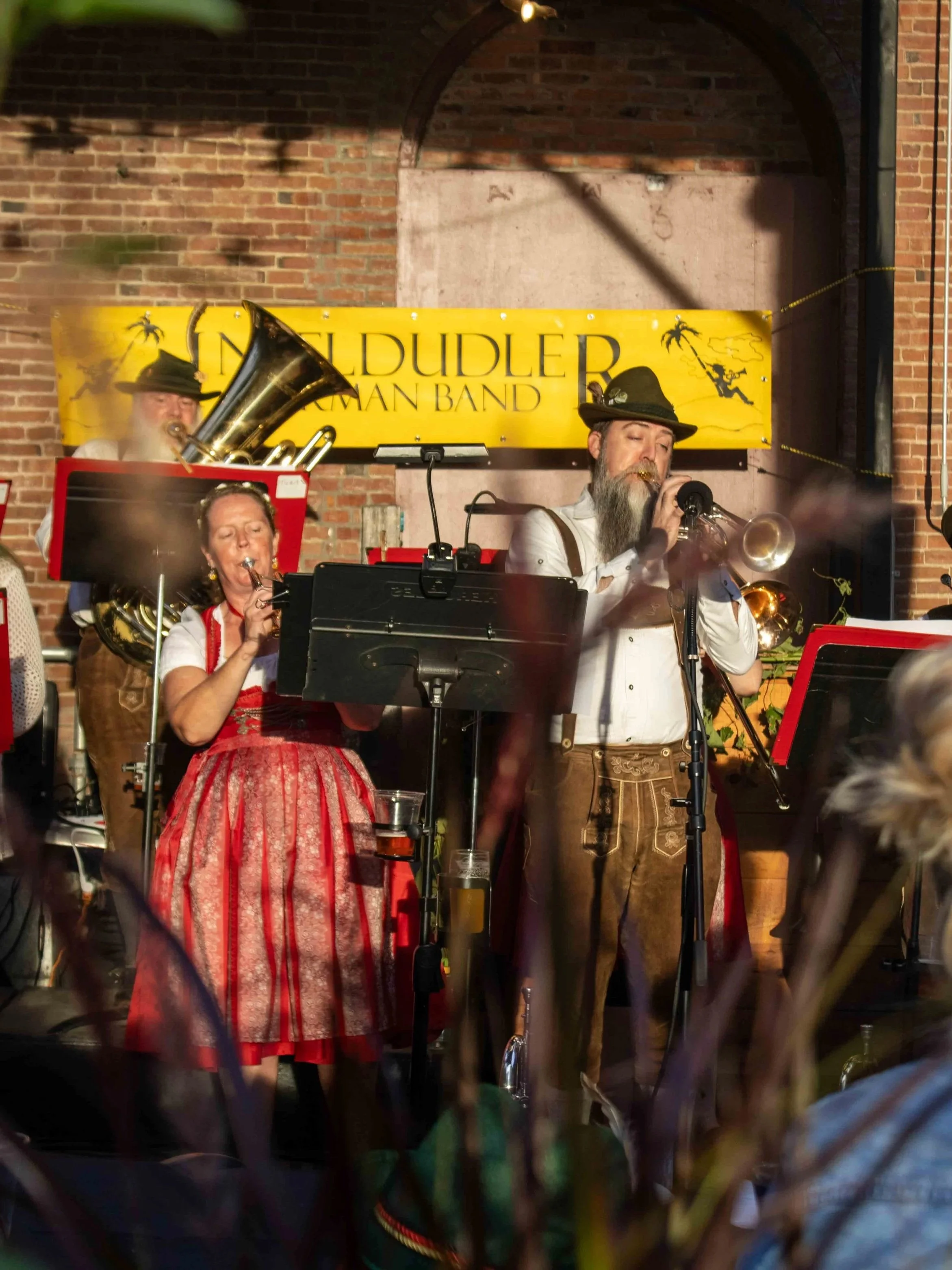 Band members dressed in traditional German attire performing with brass instruments on stage in front of a yellow banner that reads "INSELDUDLER GERMAN BAND" against a brick wall backdrop.