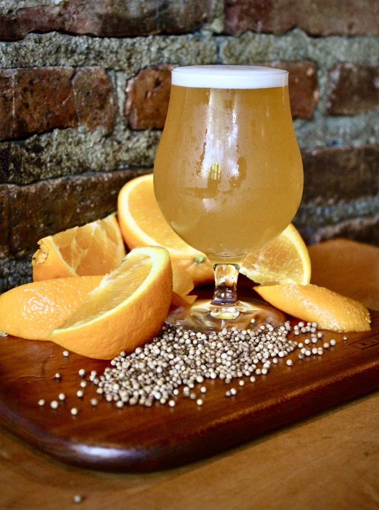 A glass of beer with a foamy head, surrounded by orange slices and white peppercorns on a wooden board, against a brick wall background.