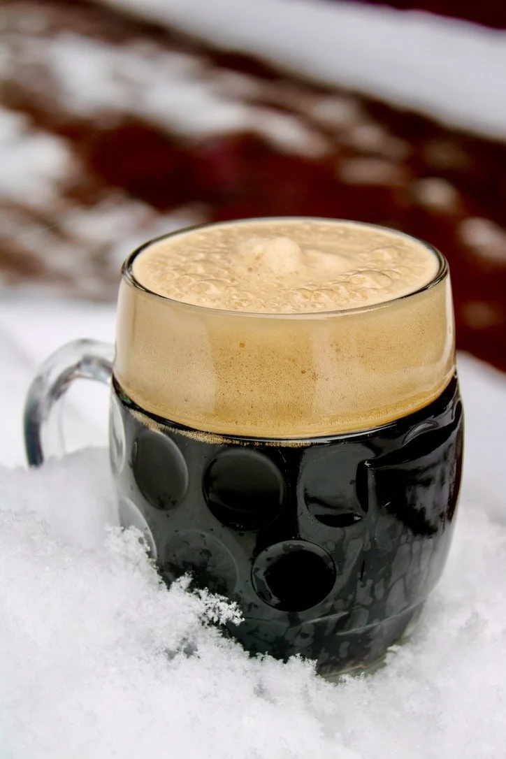 A glass mug with a black textured pattern filled with a frothy, creamy beverage, sitting on snow with a blurred background.