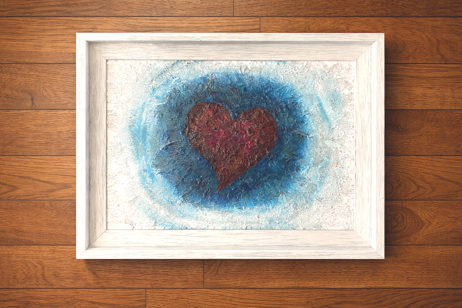 Painted artwork of a red heart with a textured surface on a blue and white background, framed in white, resting on a wooden surface.
