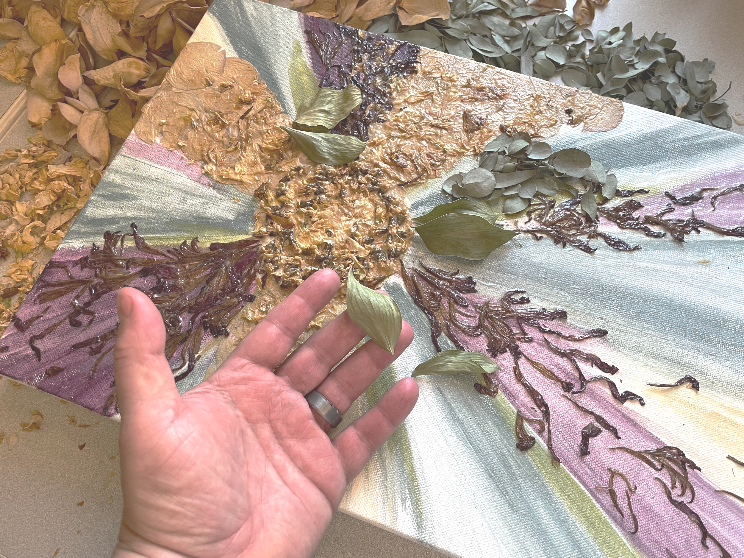 A person is holding a green leaf in front of a mixed media artwork featuring textured leaves and painted branches on a canvas.