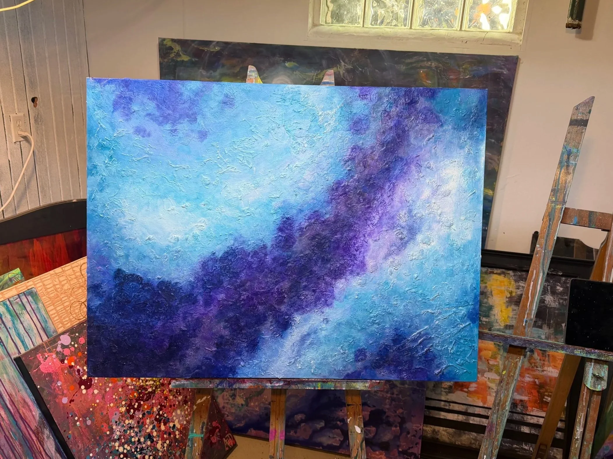 Multiple vibrant abstract paintings displayed on wooden easels inside a room with a window and a wooden-paneled wall.