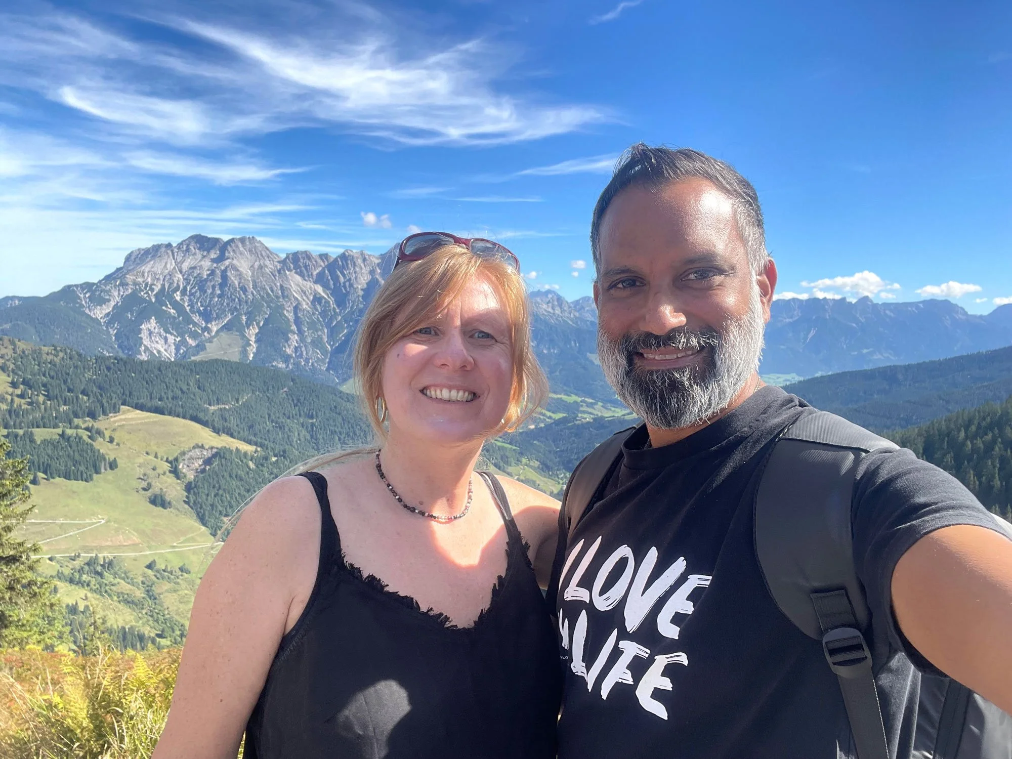 Sara and Niraj posing with mountains, green hills, and a blue sky in the background.