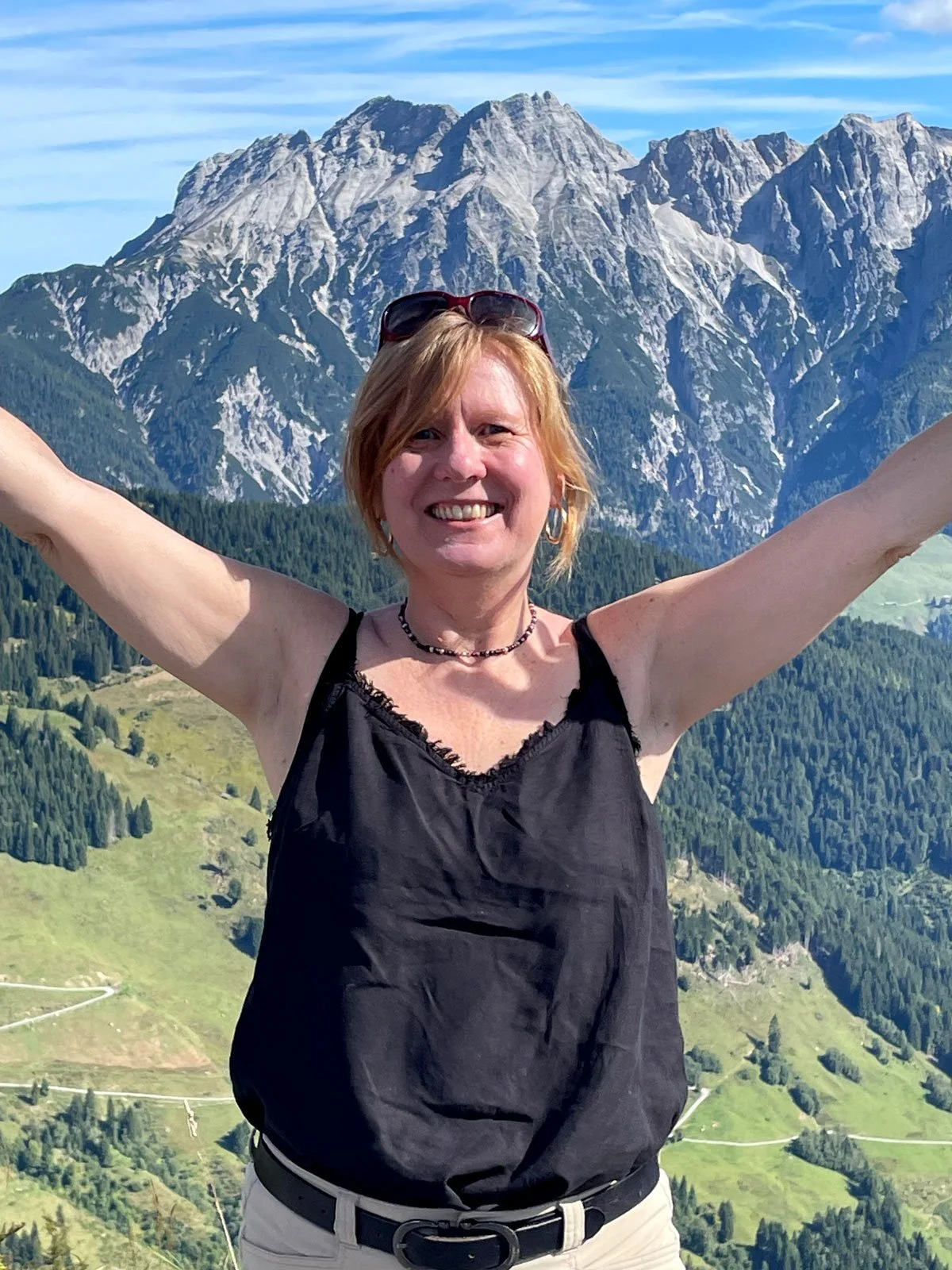 Sara smiling with arms outstretched in front of a mountain landscape with green hills and rocky peaks.