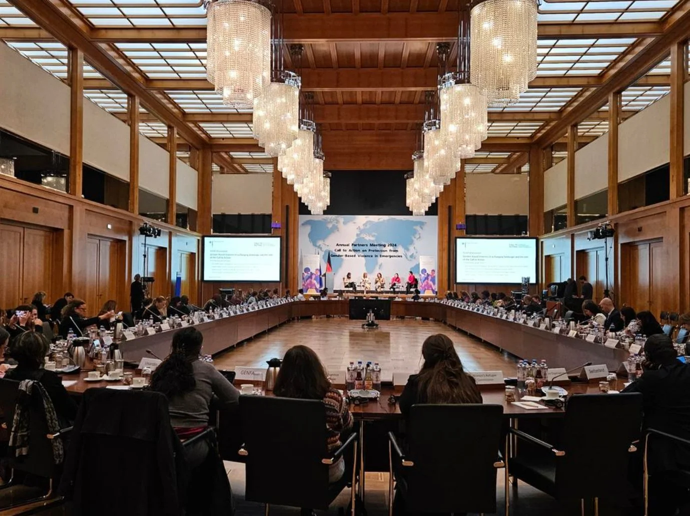 A large conference room with wooden walls and a high ceiling illuminated by a series of elegant chandeliers. Attendees are seated around a U-shaped arrangement of tables facing a stage with large screens displaying meeting information. The stage backdrop features a world map and the event title, indicating an international event.
