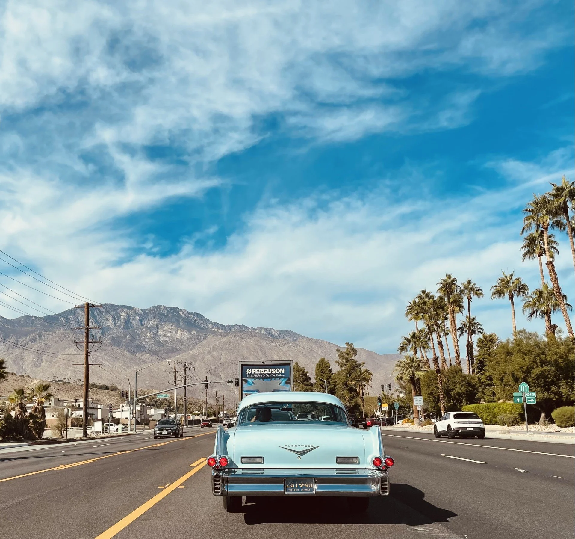 A classic light blue vintage Fleetwood car driving on a multi-lane road with palm trees, power lines, mountains, and a blue sky with wispy clouds in the background.