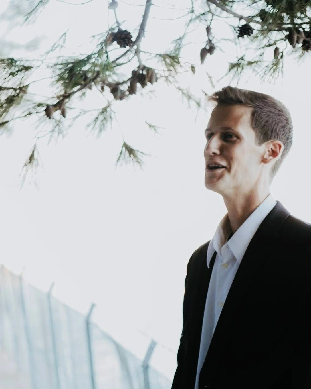 A man in a black suit and white shirt talking outdoors near tree branches and pinecones.