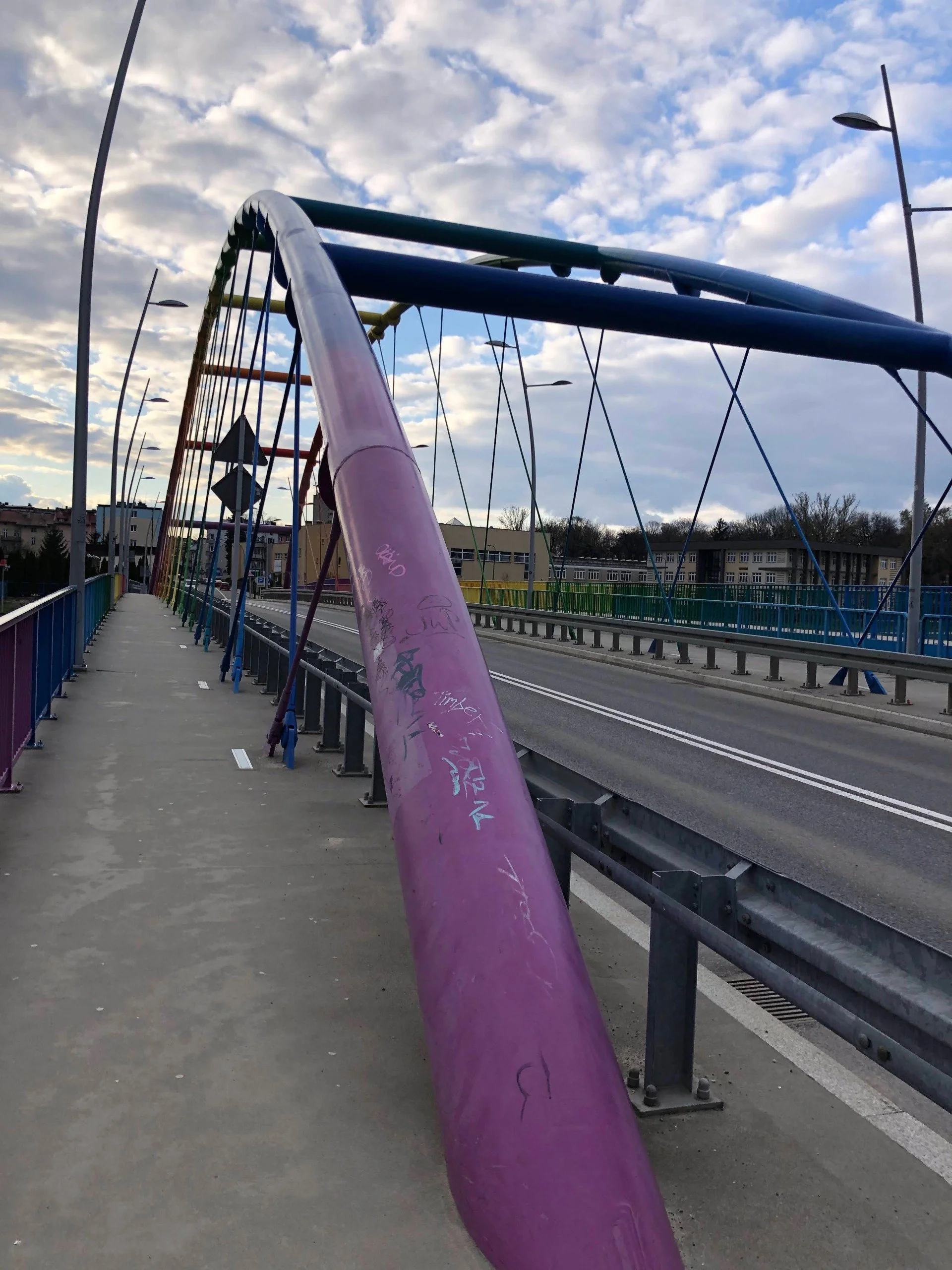 Sidewalk on a bridge with graffiti-covered purple and multicolored railings, over an empty road, under cloudy sky.