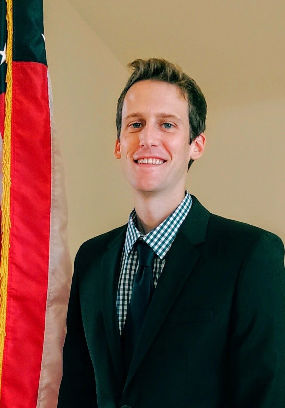 A smiling young man in a dark suit with a checkered shirt and tie, standing next to a flag with red, black, and beige colors.