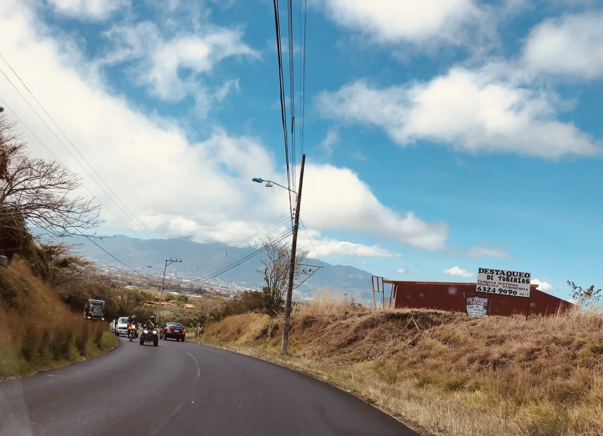 A winding rural road with several cars and motorcycles, dry grass and trees on the sides, utility poles with power lines, mountains in the background, and a sign advertising pipe repairs and sales.