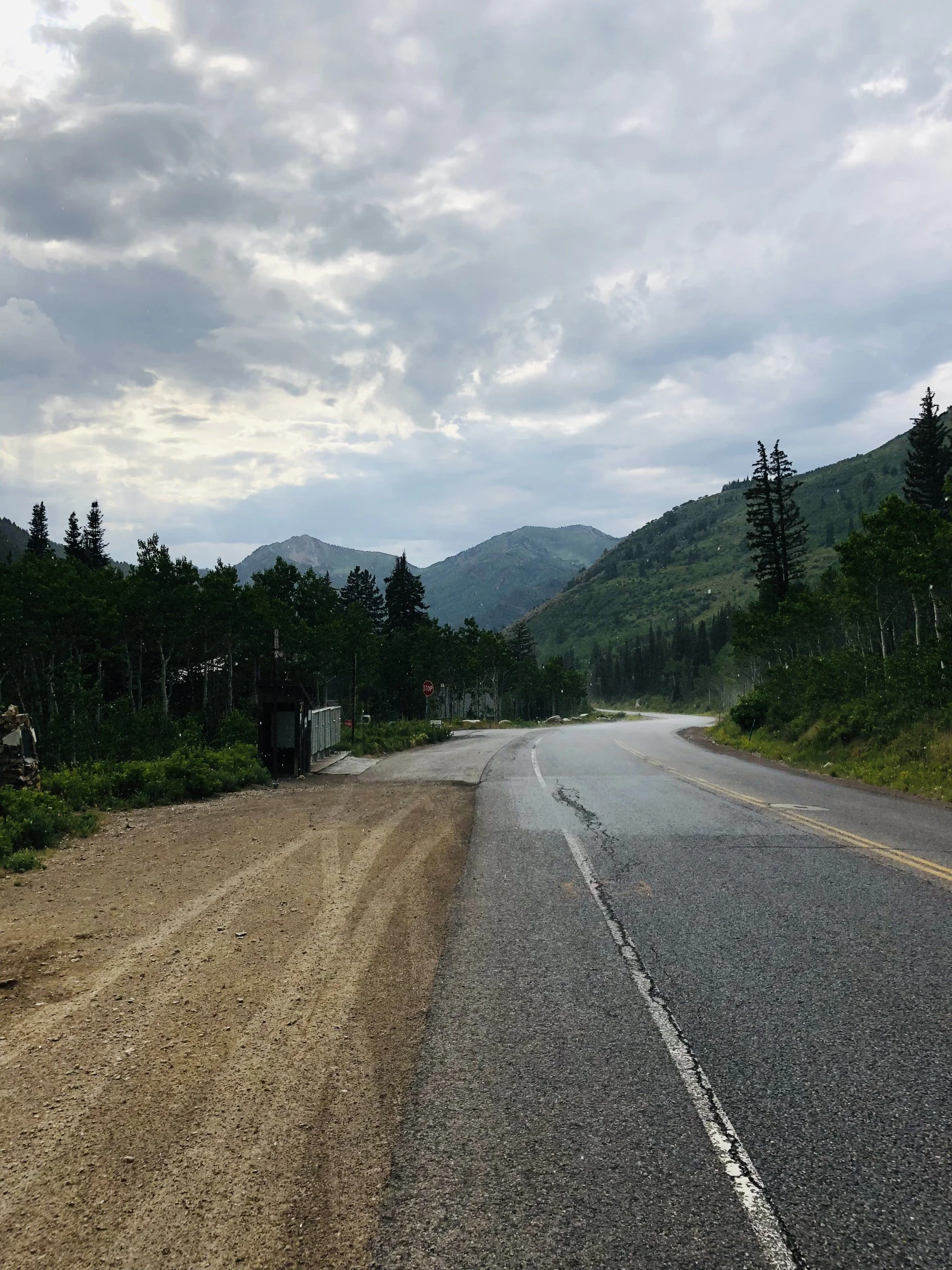 A winding mountain road with a dirt shoulder on the left, surrounded by green trees and mountains under a cloudy sky.