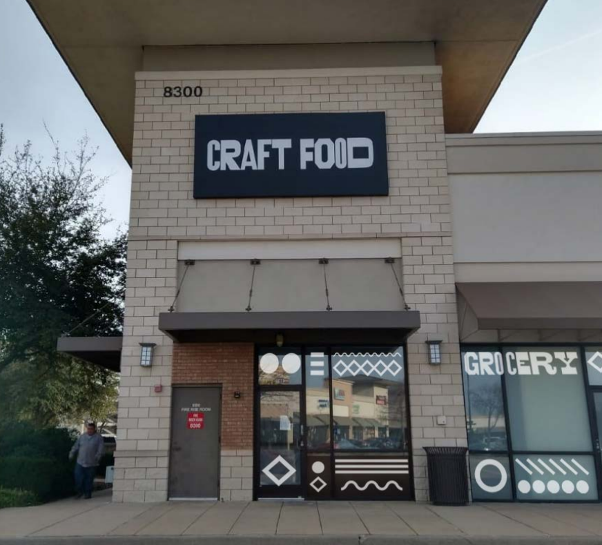 Store front of Craft Food with a large sign, brick and white exterior, large glass windows, and abstract graphics on the door and windows.