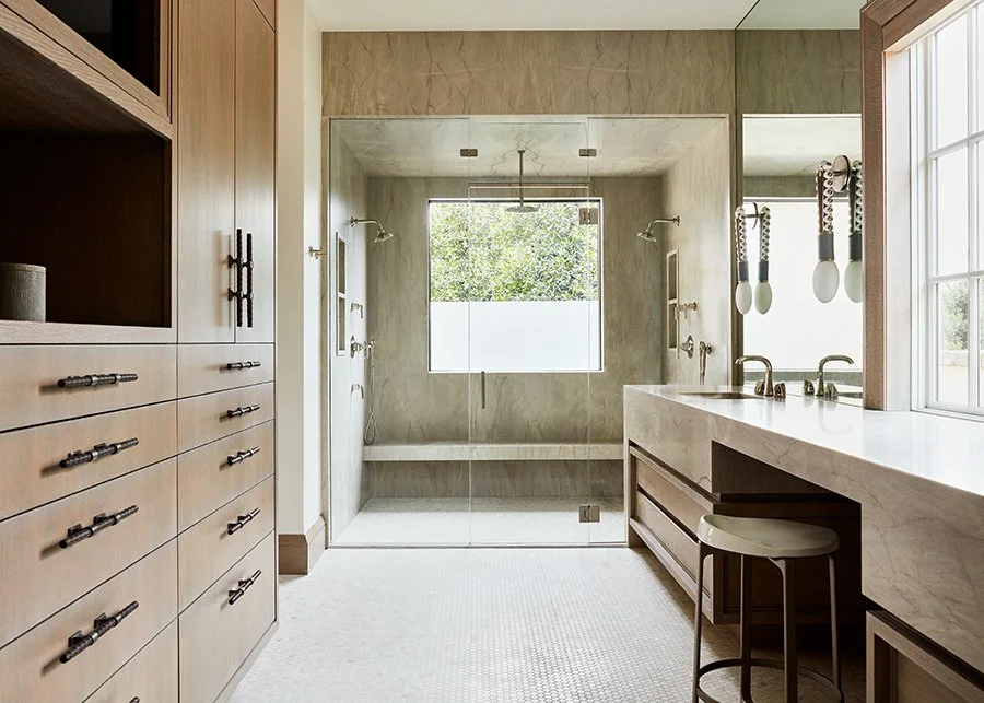 Modern bathroom with a walk-in glass shower, wooden cabinetry, marble counters, and a large window overlooking trees.