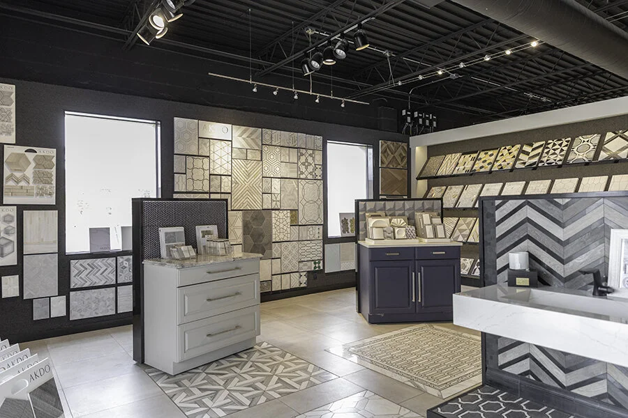 Interior of a tile and material showroom with displays of various tile samples on walls and shelves, featuring different patterns and colors, with a reception desk and displayed sample boards.