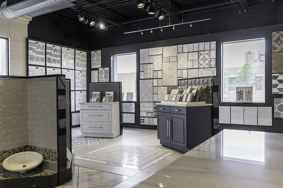 Showroom displaying various tile and backsplash samples, with display boards on the walls, a black cabinet with tile samples, and a tile display with a mosaic sink in the foreground.