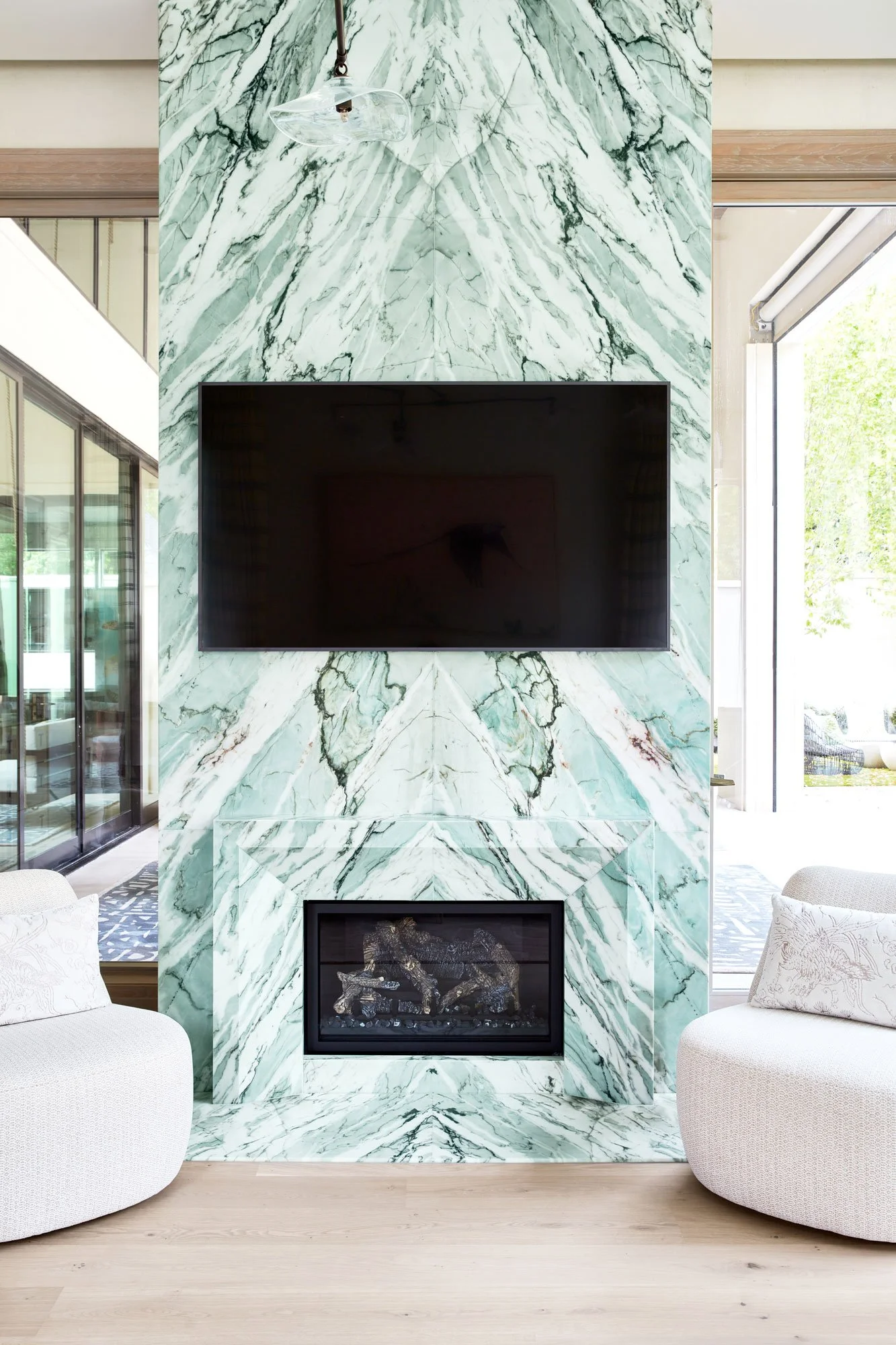 Living room with a green and white marble fireplace, a flat-screen TV mounted above it, two white armchairs on either side, and large glass doors letting in natural light.