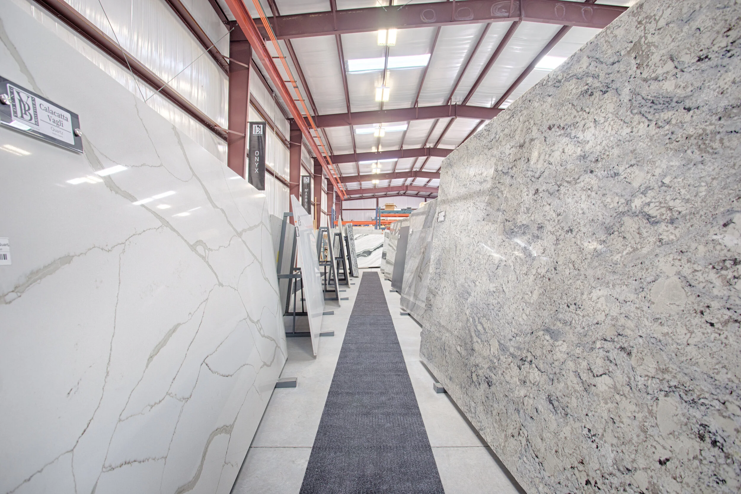 A warehouse with large slabs of marble and granite stone on display, arranged on metal stands along a carpeted walkway. The warehouse has a high ceiling with exposed beams and skylights.