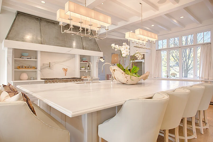 Bright kitchen with white island, white chairs, floral centerpiece, large windows, and modern lighting fixtures.