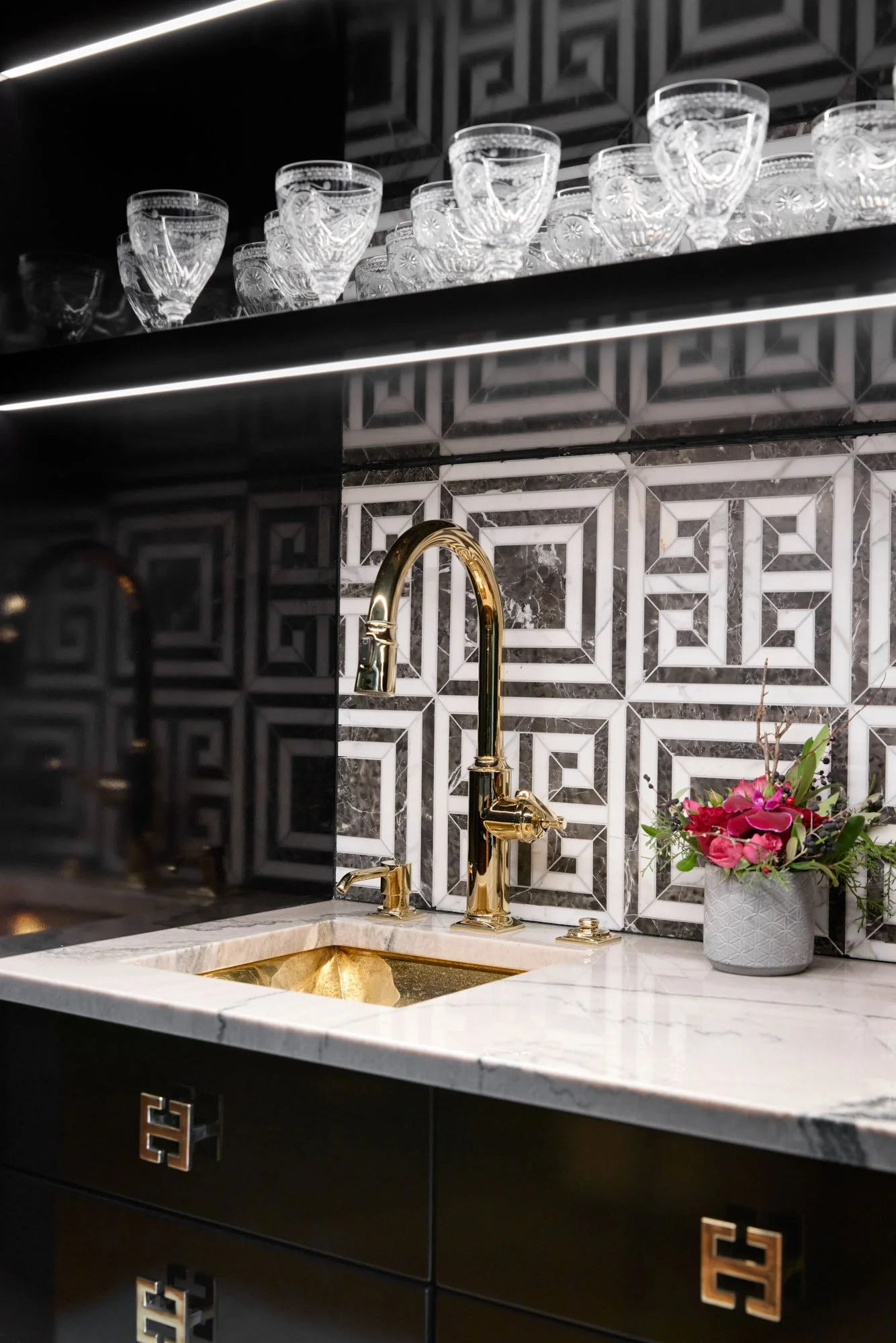 A kitchen with black cabinetry featuring geometric gold handles, a marble countertop, a gold faucet, a marble backsplash with black, white, and gray geometric patterns, a shelf with crystal glasses, and a gray vase with pink flowers.