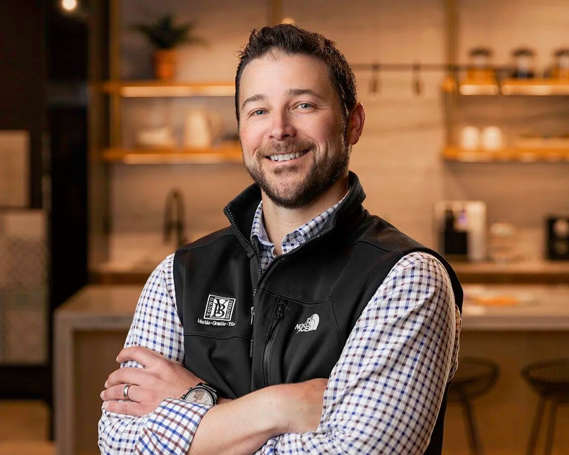 A smiling man with a beard and short dark hair, wearing a plaid shirt and a black vest, standing with arms crossed in a modern, warmly lit space with wooden shelves and barstools in the background.